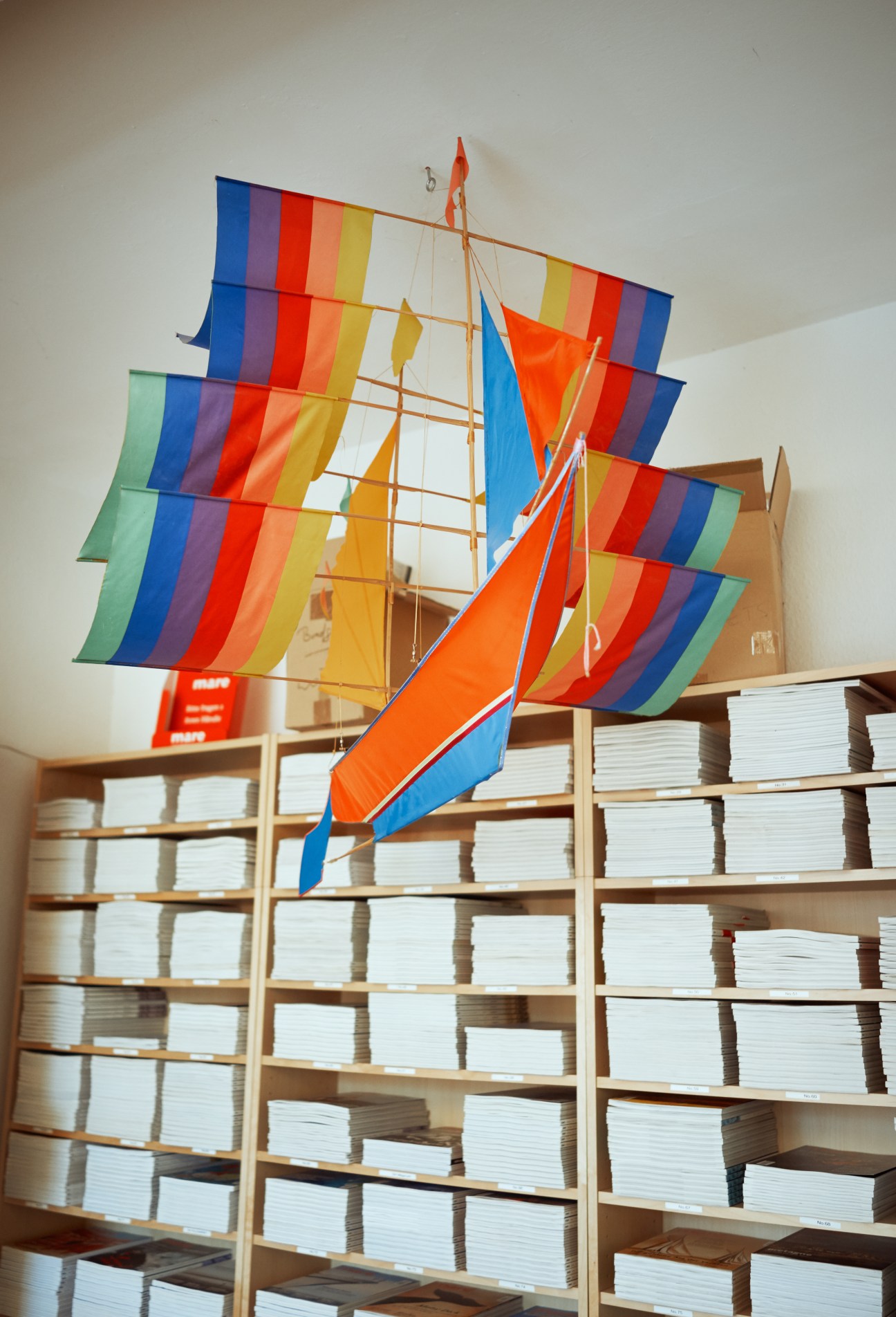 Colorful toy sailboat with rainbow-striped sails displayed above wooden shelves stacked with white magazines.