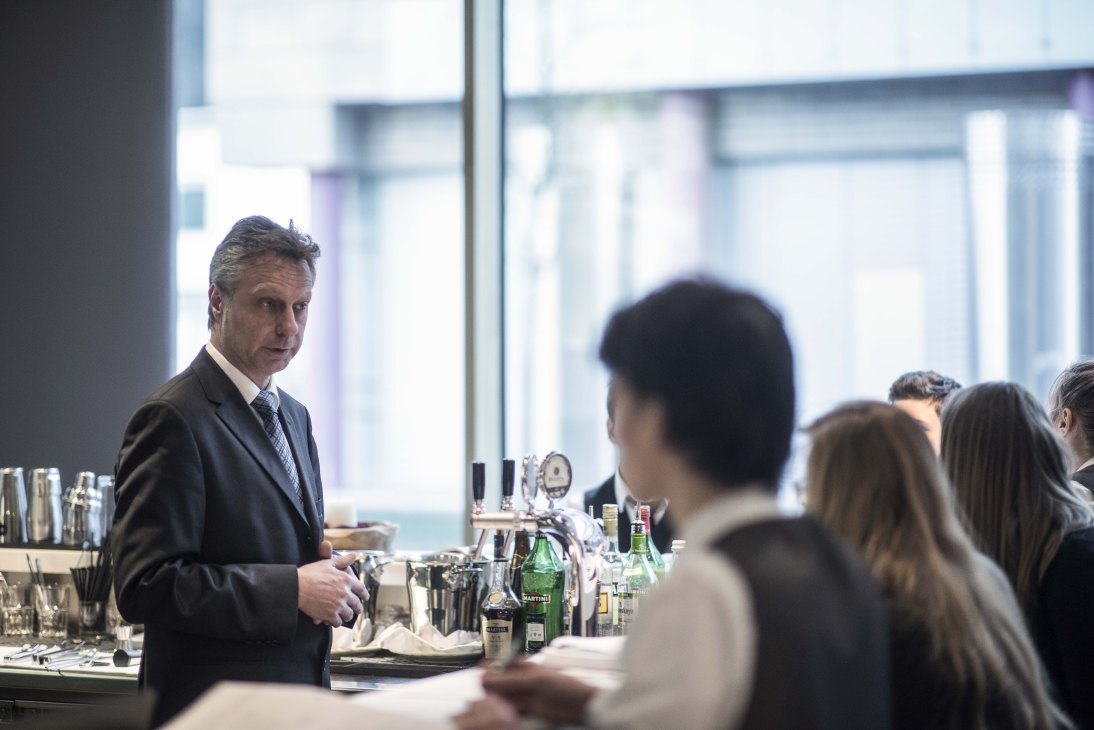 A Brunico Hotel School instructor in a dark suit gives a bartending lesson to students at a bar counter with bottles and cocktail equipment.