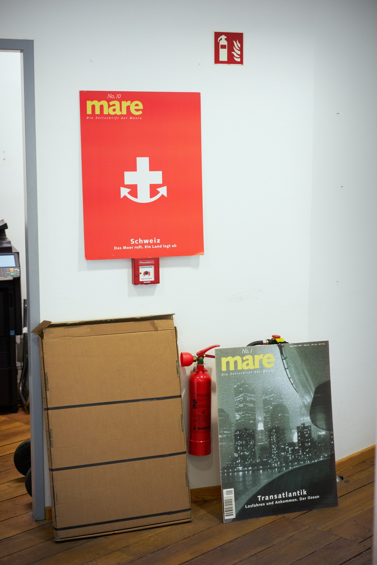Mare magazine cover art displayed on a white wall above a cardboard box and red fire extinguisher in an office setting.