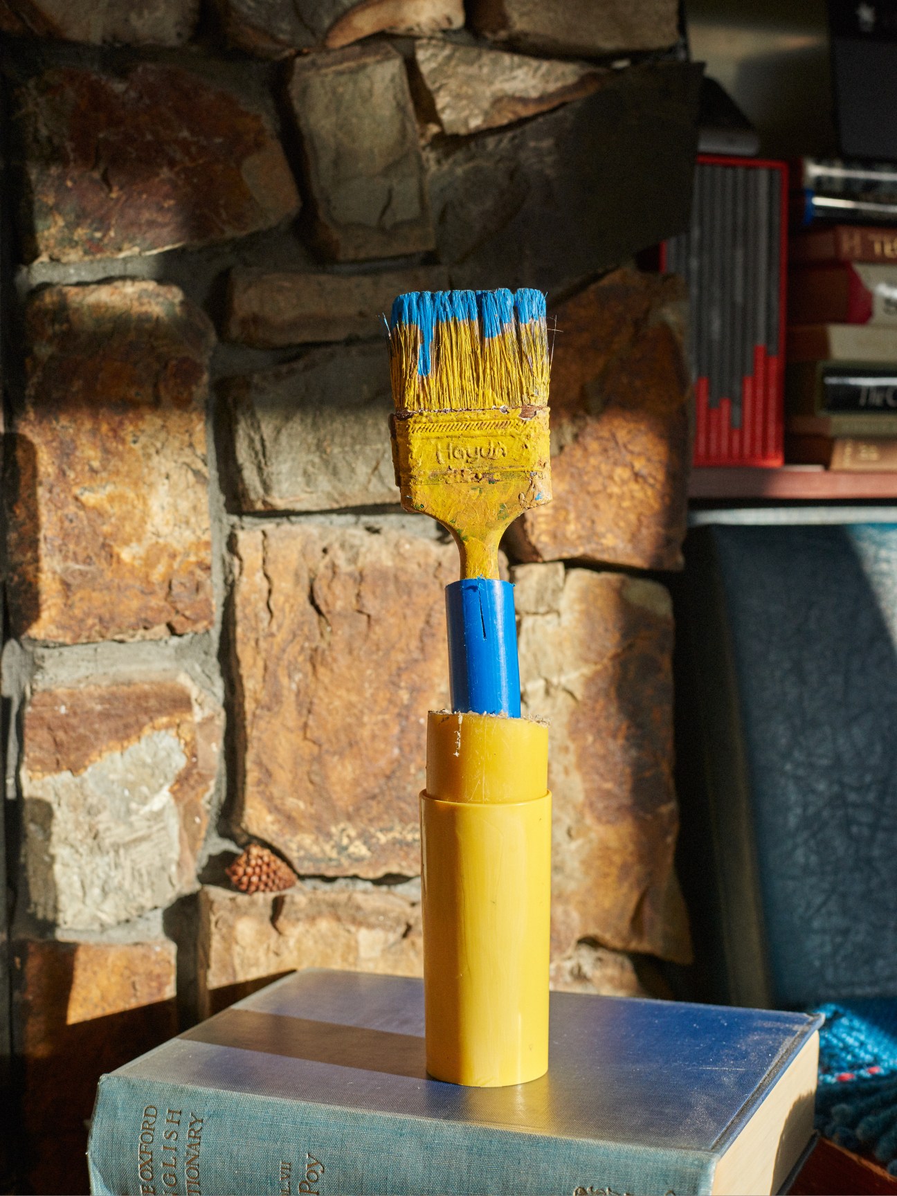 Sculpture by New Zealand artist Don Driver featuring a paintbrush with blue and yellow painted handle against a stone wall.