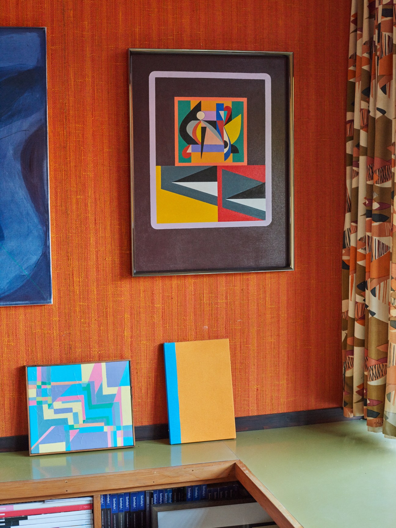Framed abstract painting by Diena Georgetti displayed on orange textured wall with colorful geometric artwork nearby.