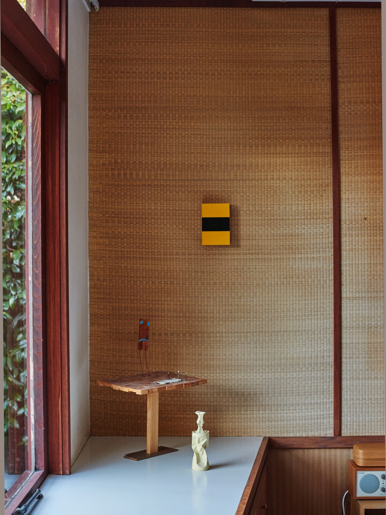 Master bedroom with tatami-covered walls, wooden pedestal table, geometric yellow and black artwork, and small sculptural object.