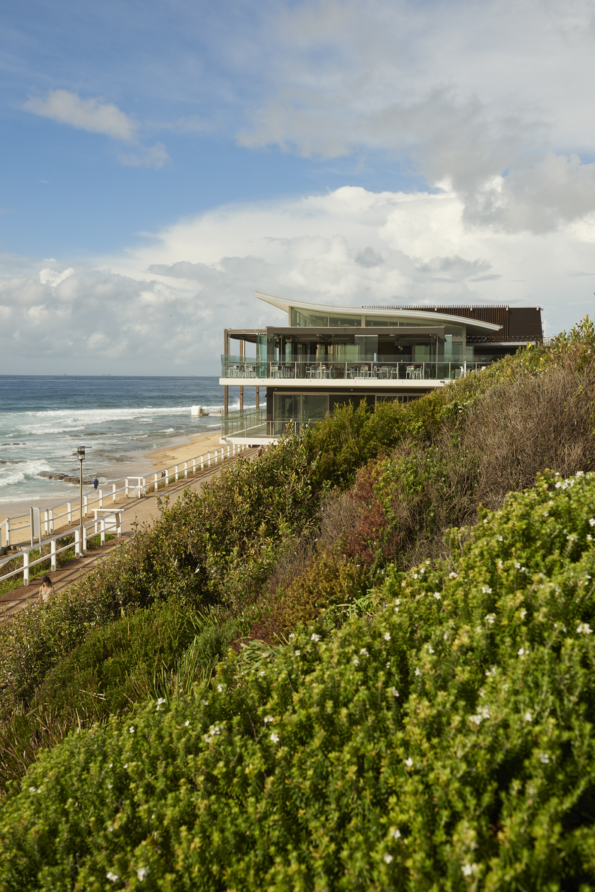 Modern beachfront building with glass panels and angled roof overlooking sandy beach and ocean waves.