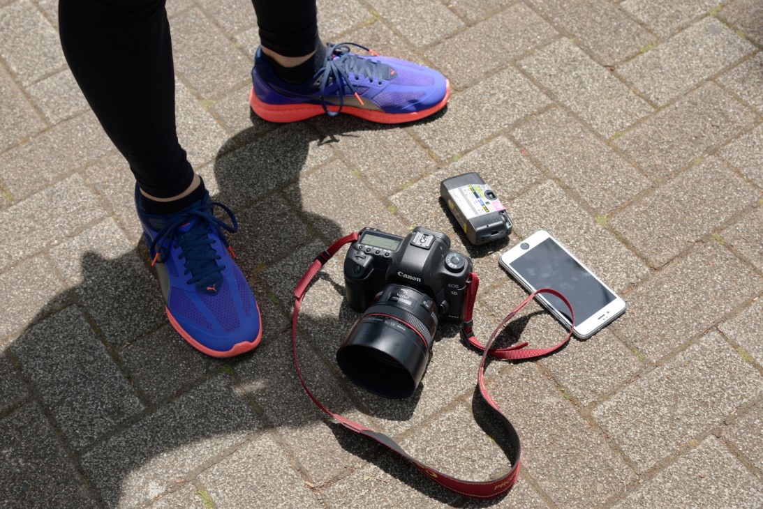 Runner wearing blue and coral shoes stands next to photography equipment including a Canon camera, smartphone, and flash unit on pavement.