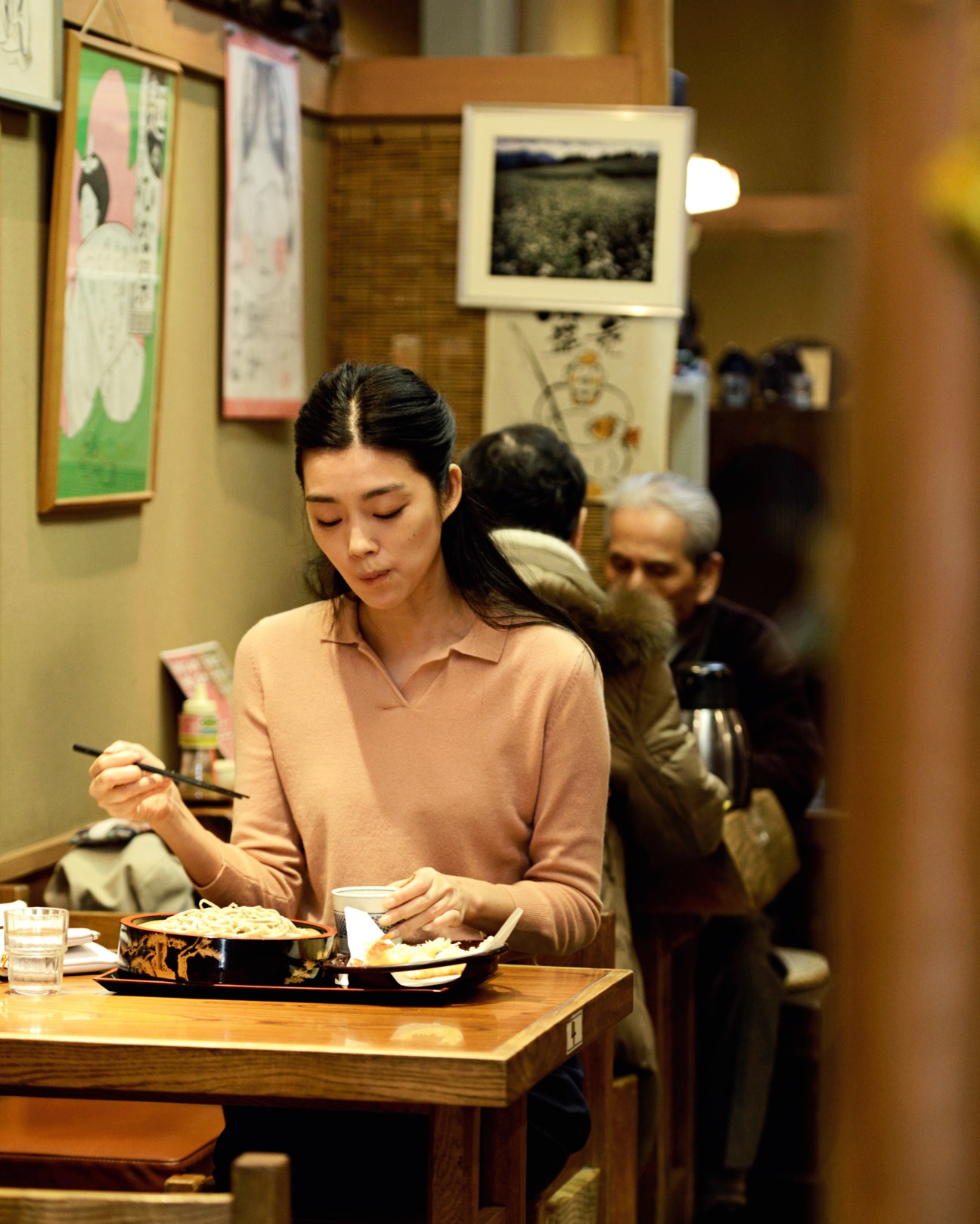 Woman in peach polo shirt eating cold soba noodles with chopsticks at a Tokyo restaurant counter.