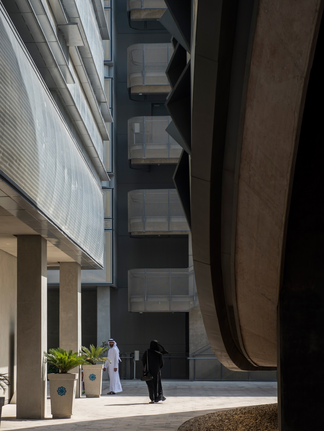 Two figures walk through a sunlit courtyard flanked by modern terracotta and glass buildings in Masdar City.