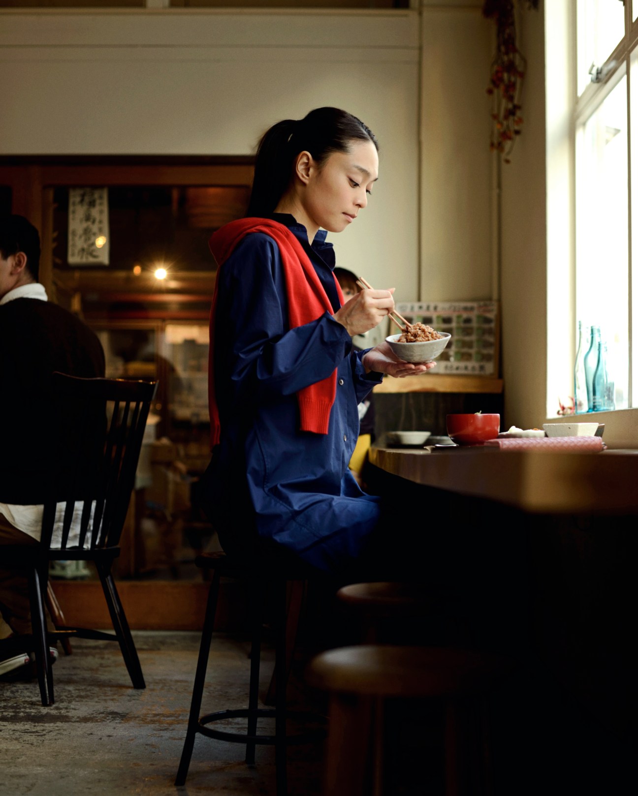 Woman in navy dress and red scarf eating noodles at a counter in a Tokyo restaurant.