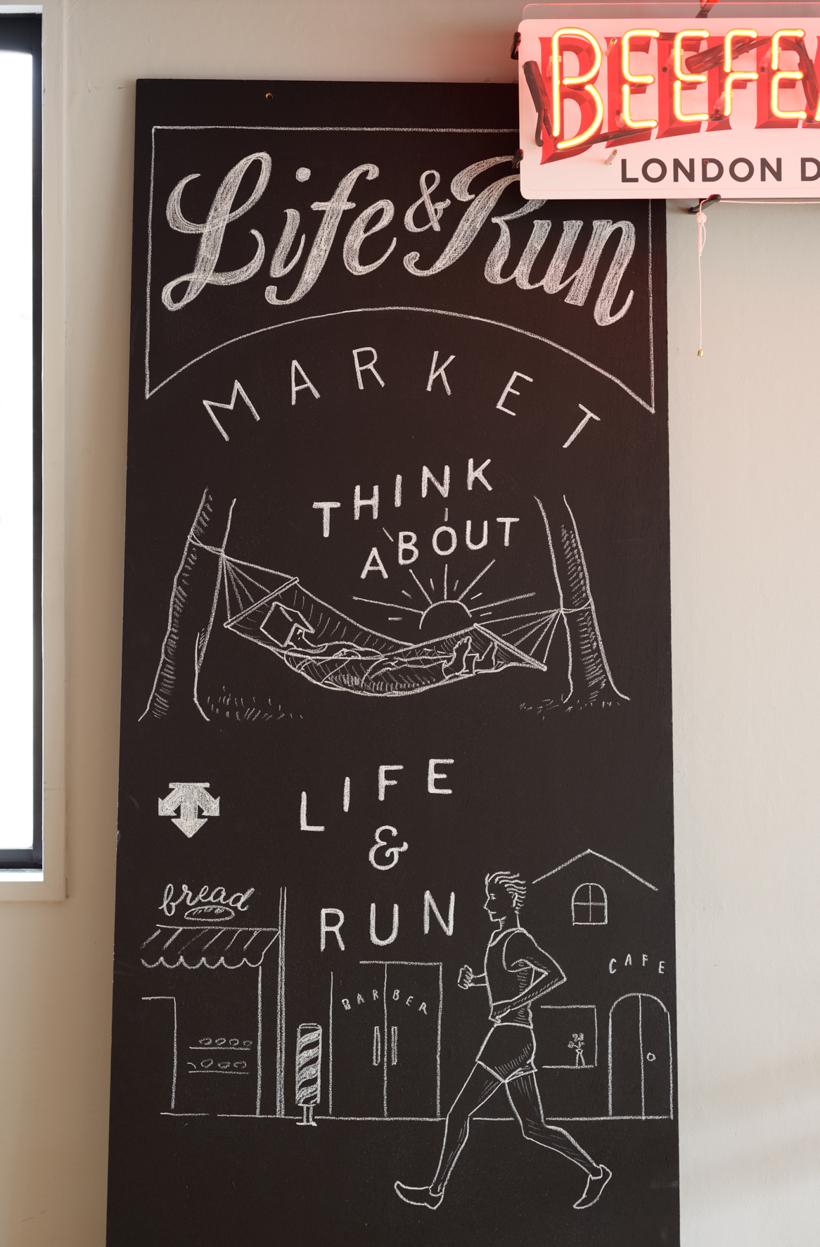 Black chalkboard sign for Life & Run Market with white hand-drawn lettering and illustrations of a runner, storefronts, and landscape.