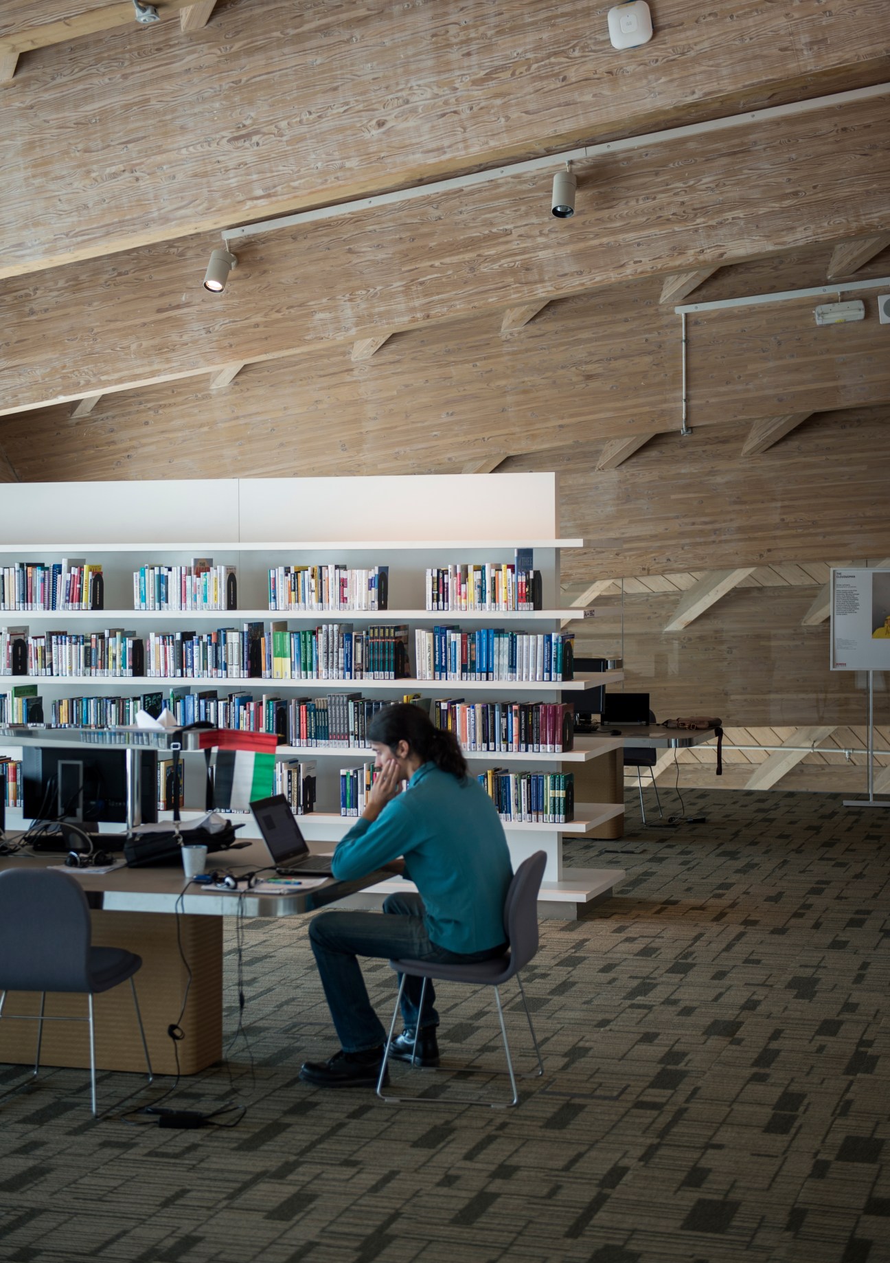 Person working at laptop in modern library with wooden ceiling and built-in bookshelves.