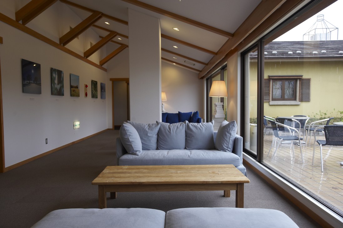 Modern hotel lounge with gray sofa, wooden table, exposed beams and large windows overlooking a terrace.