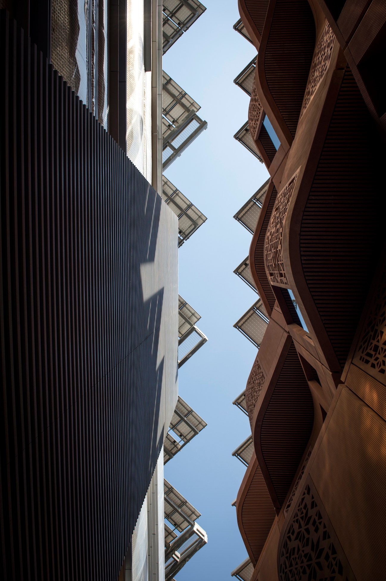 Upward view of narrow street in Masdar City with rust-colored building on right and light-colored building with solar panels on left.