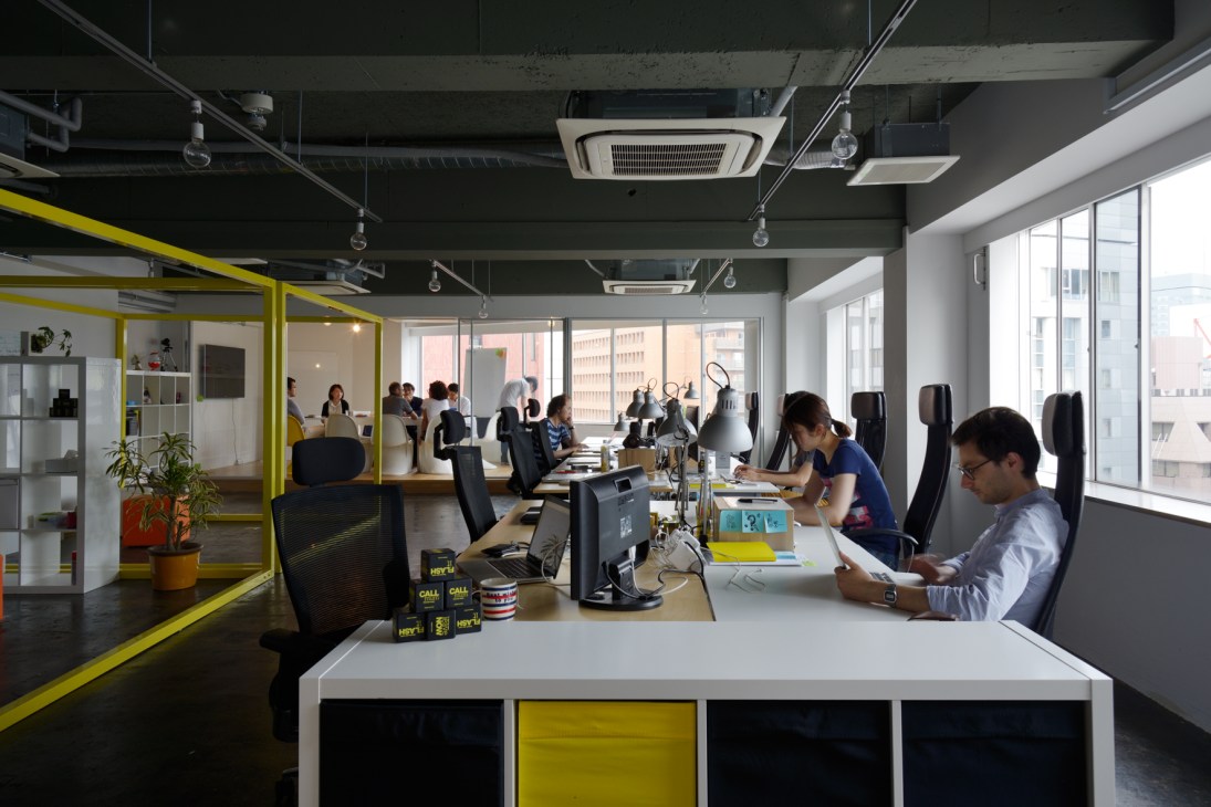 Modern office workspace with yellow-framed partitions, white desks, and large windows overlooking Tokyo buildings.