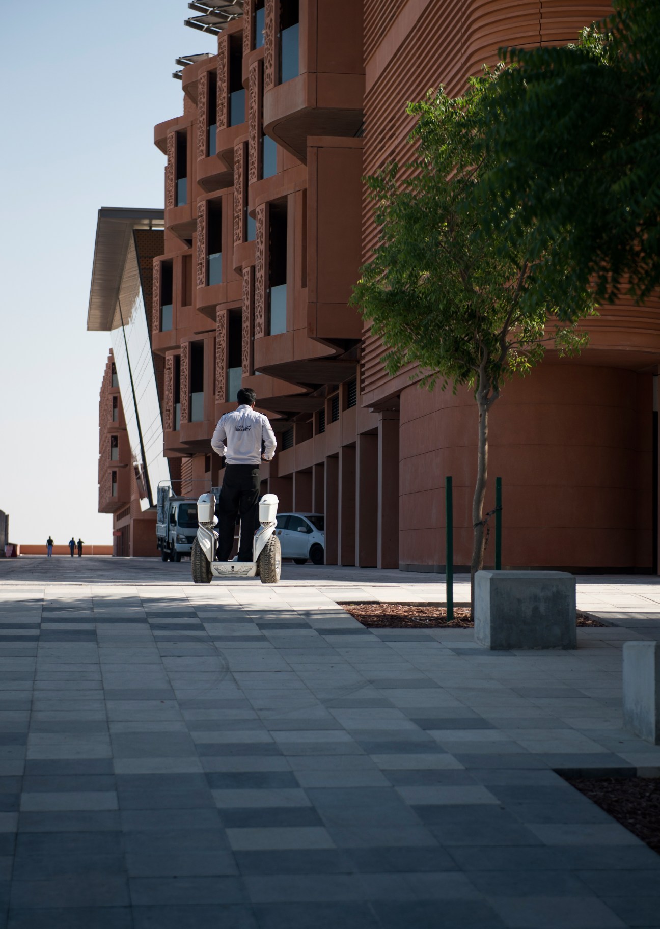 Person riding a Segway on a wide plaza in front of rust-colored modernist buildings at Masdar City.