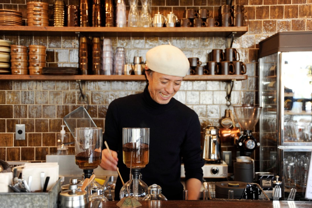 Man in white cap and black sweater operating a vacuum siphon coffee maker at Bird café counter.