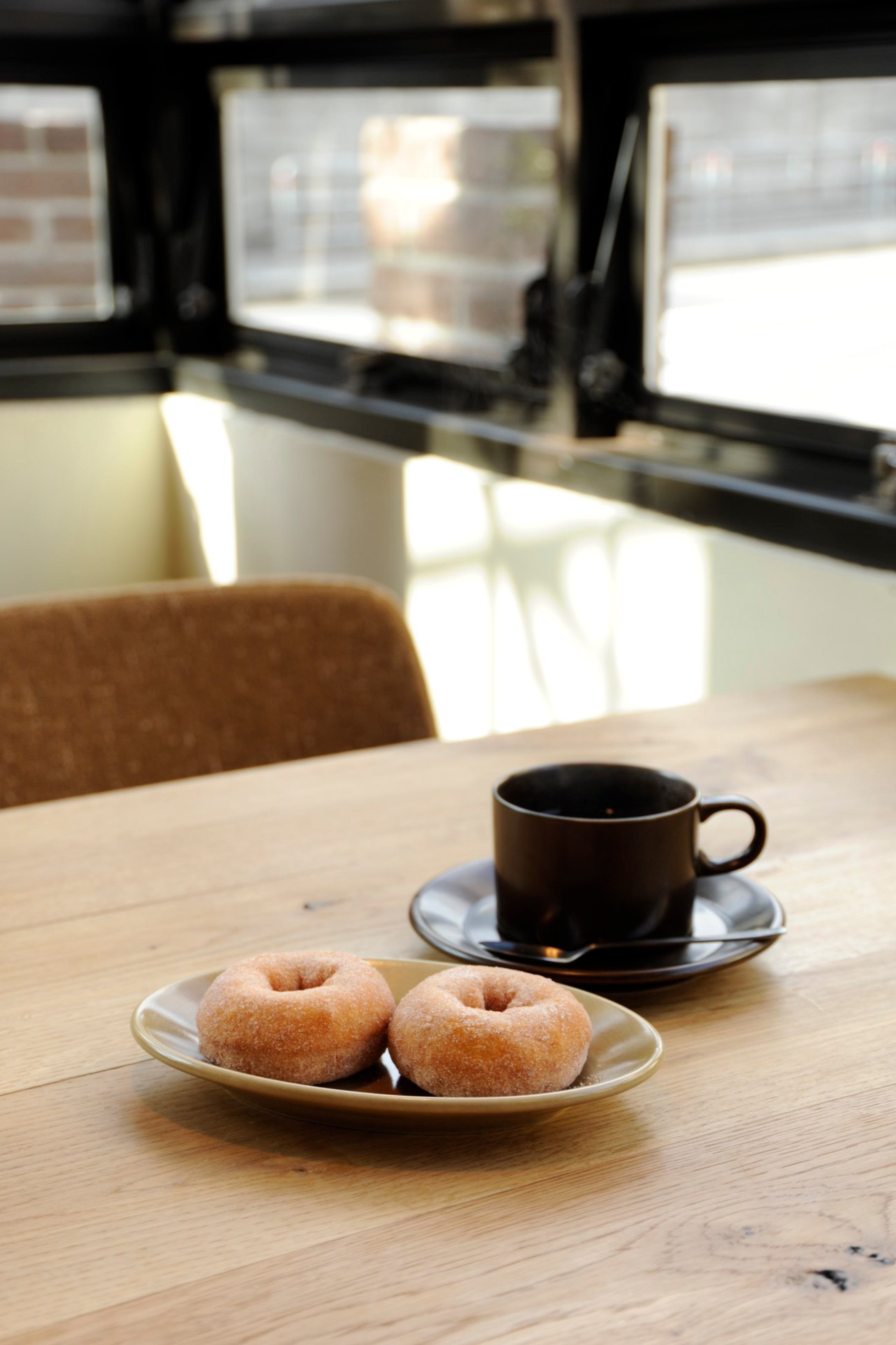 Two glazed doughnuts on a ceramic plate with a black coffee cup and saucer on a wooden table.