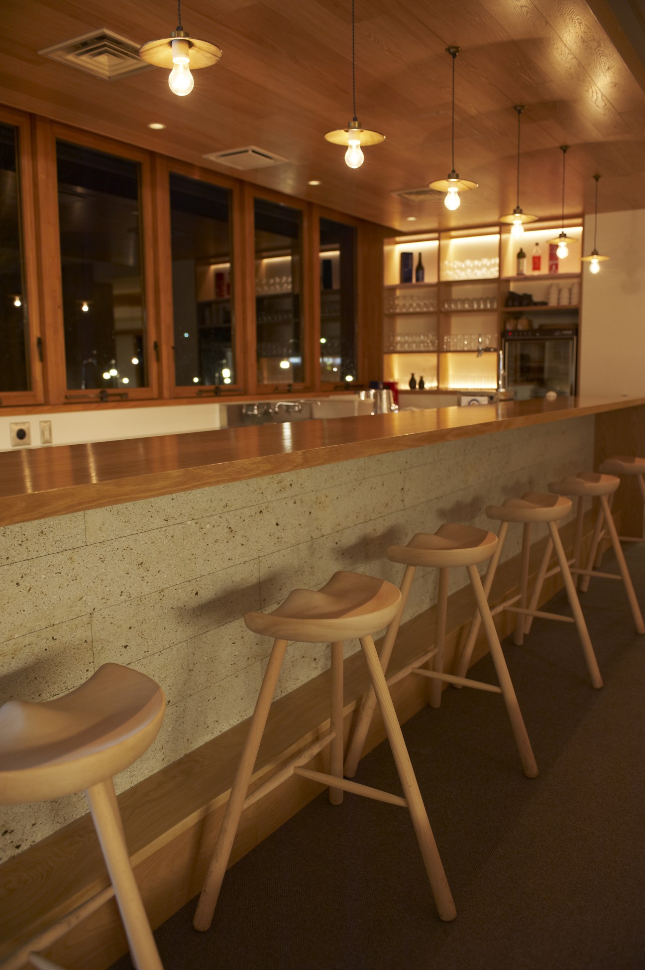 Modern hotel bar with wooden stools, pendant lighting, and illuminated shelving displaying bottles.