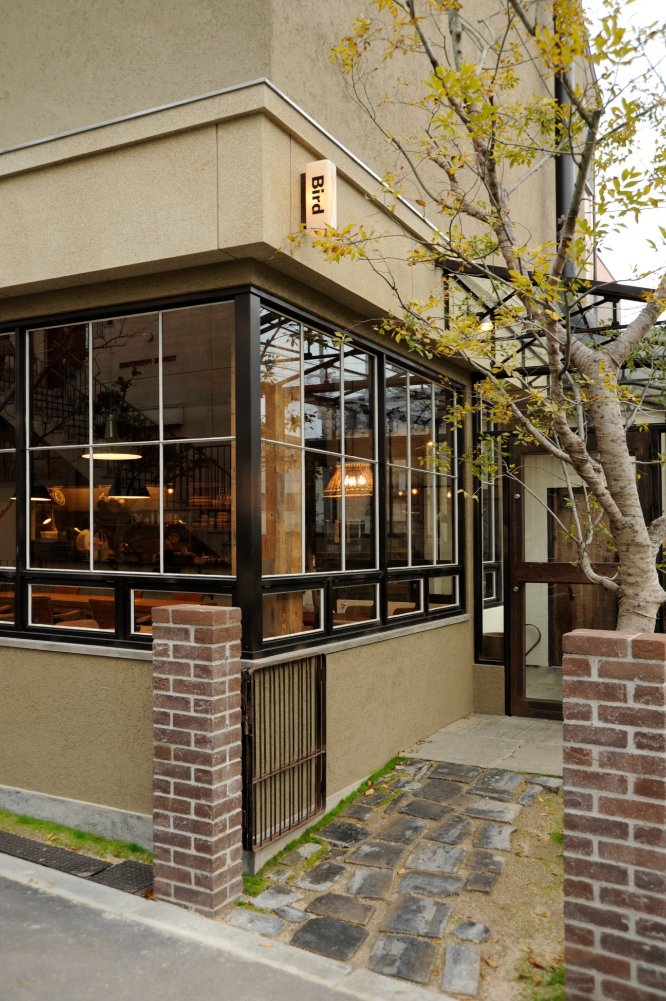 Exterior of Bird café entrance with black-framed glass windows, brick pillars, and golden tree foliage.