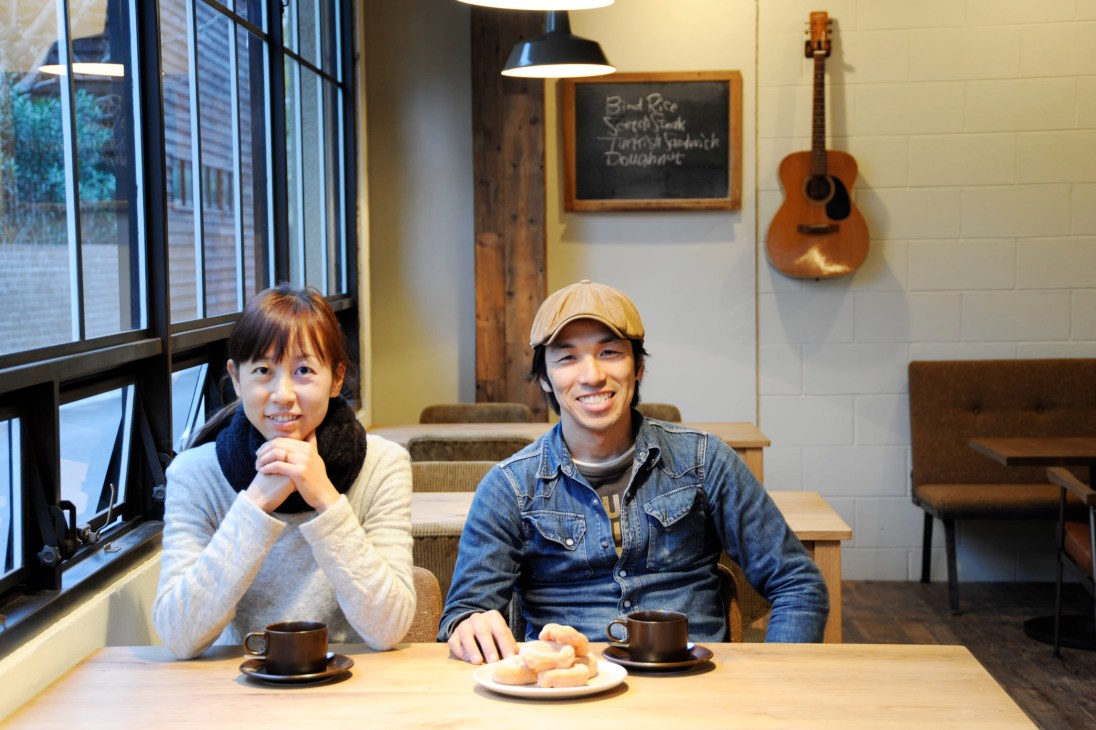 Hiromi Karatsu and Tokuhiko Kise sit at a table in their Bird café, smiling at the camera with coffee cups and doughnuts.