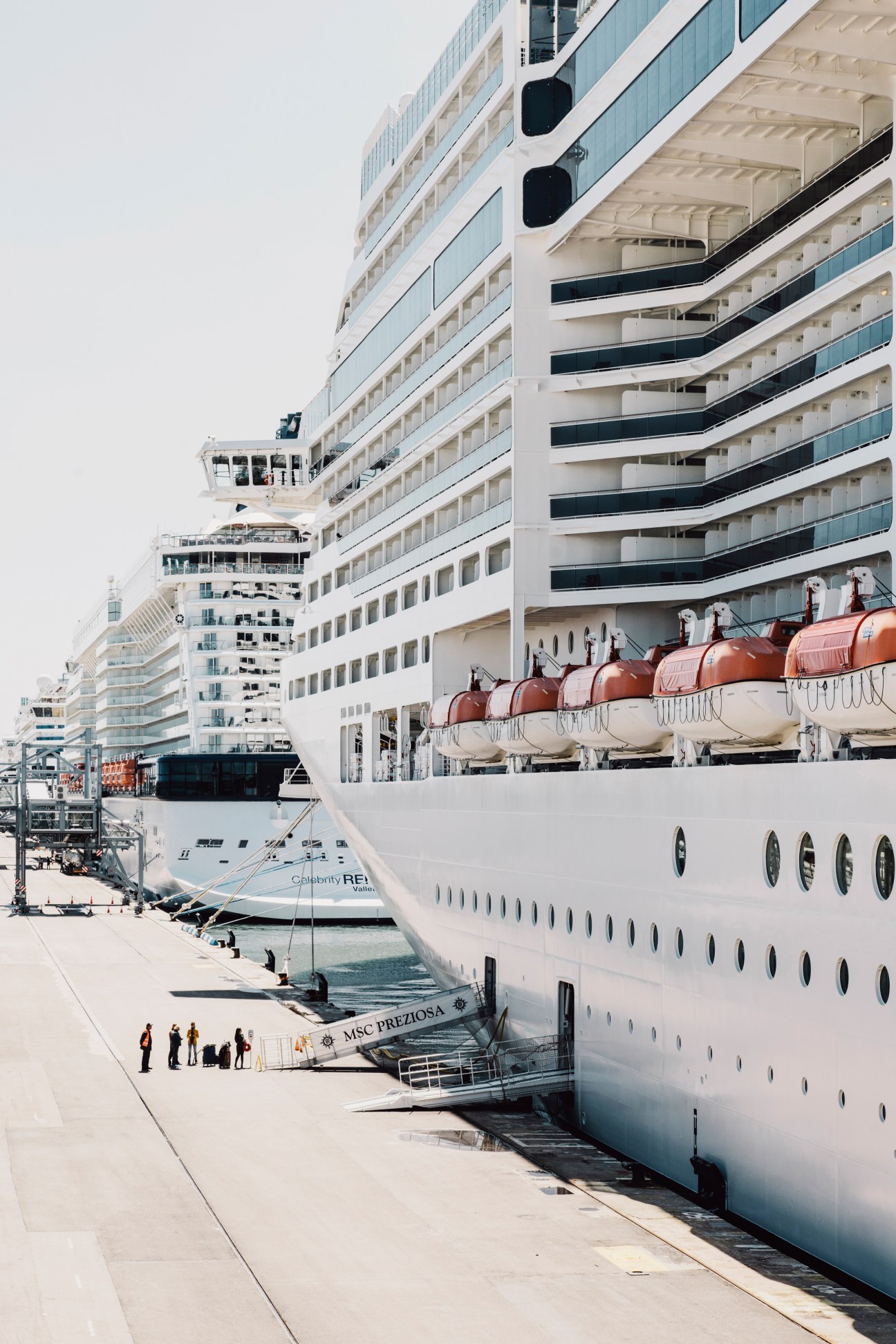 MSC Preziosa cruise ship docked at port with orange lifeboats visible on deck and other vessels nearby.