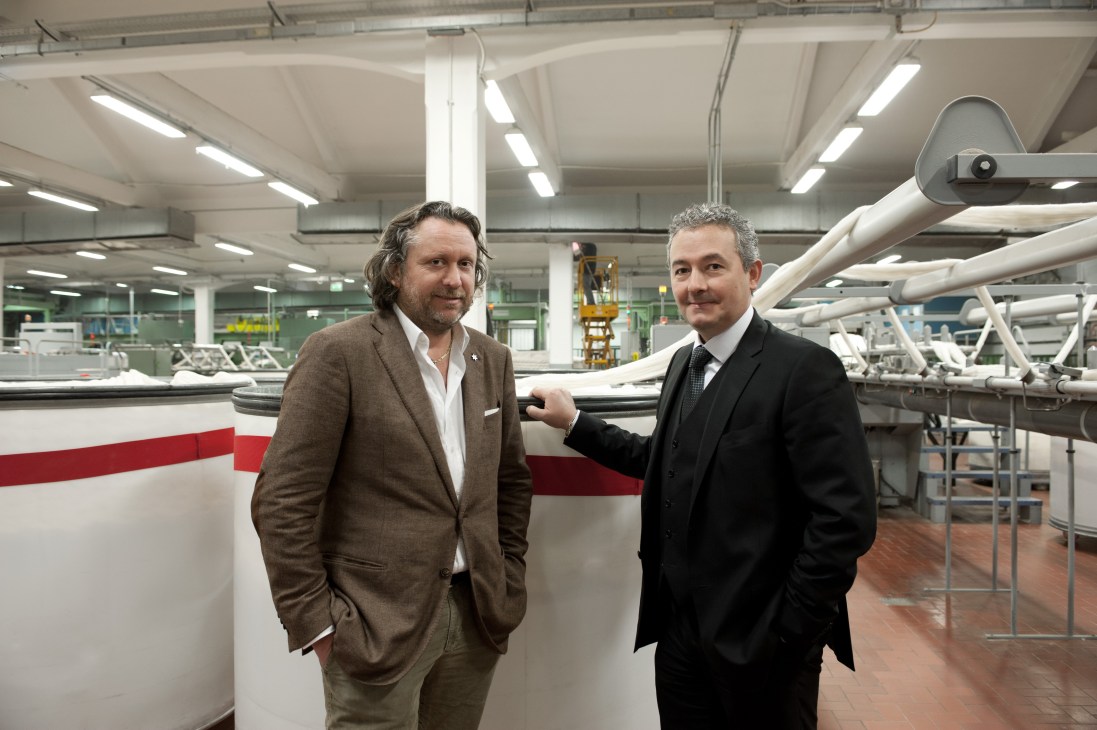 Two men standing in a textile mill with industrial machinery and large white fabric rolls visible in the background.