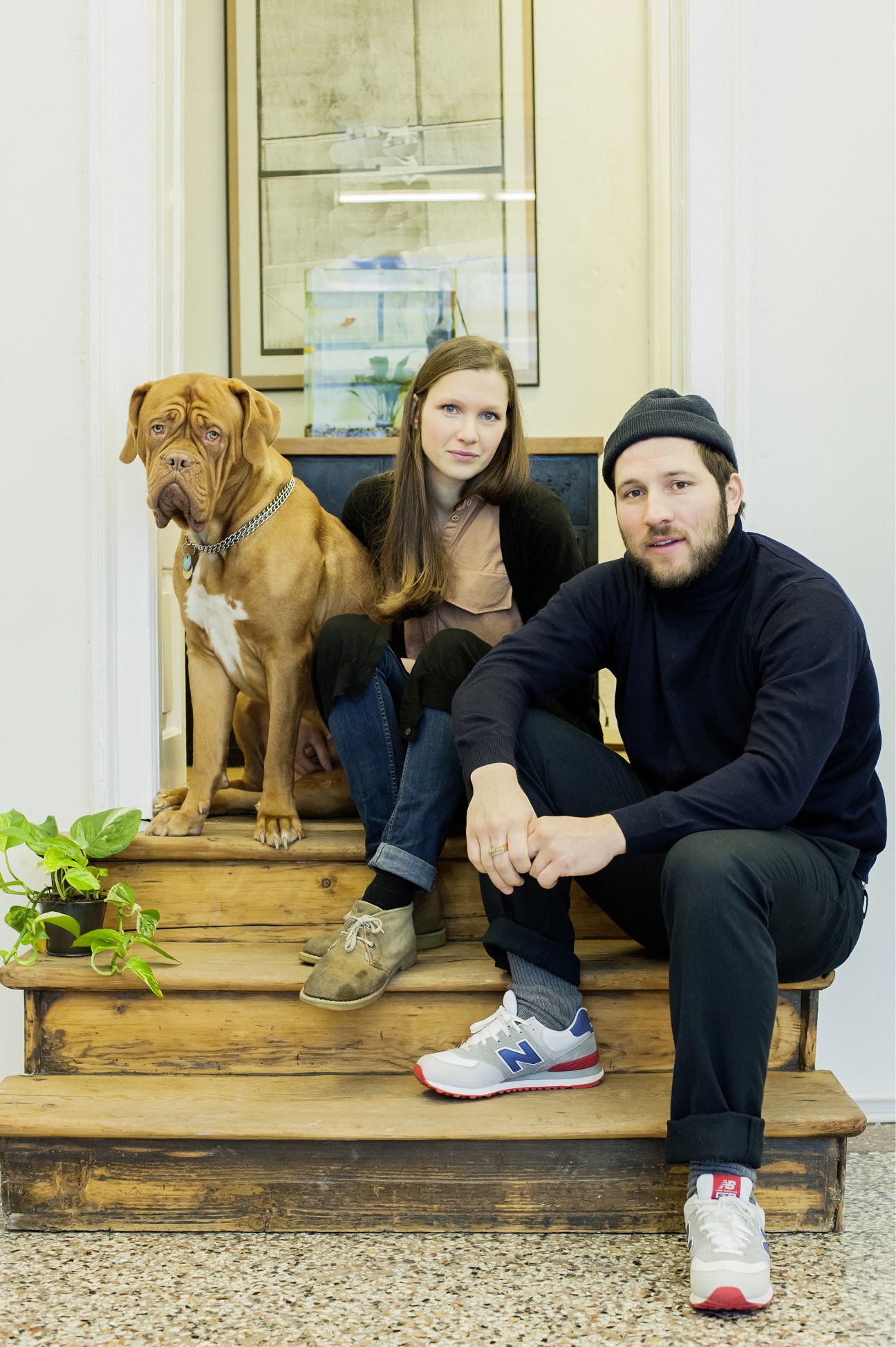 Two people and a brown dog sit on wooden steps in a minimalist interior space.