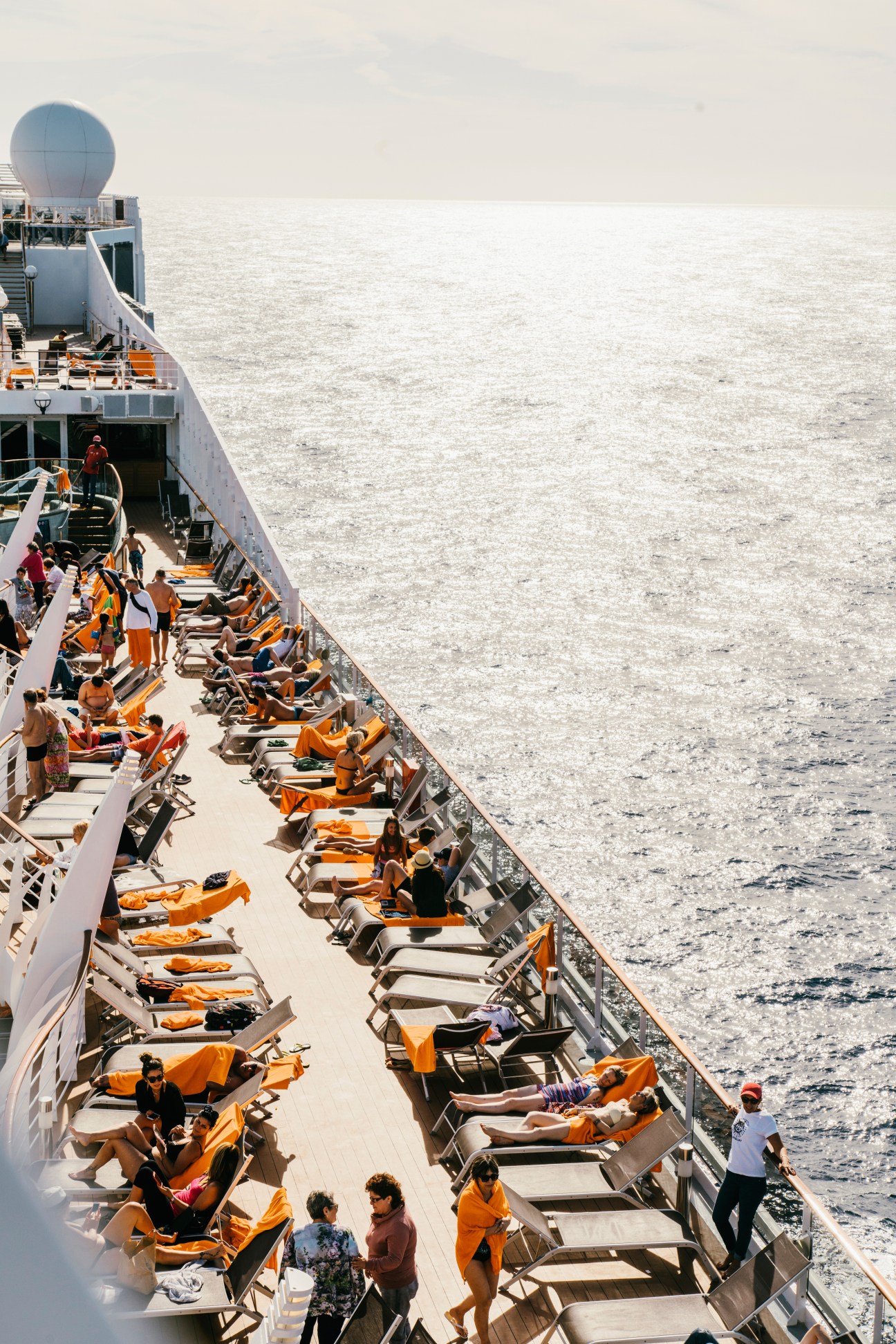Cruise ship deck with rows of orange sun loungers and passengers relaxing on the open water.