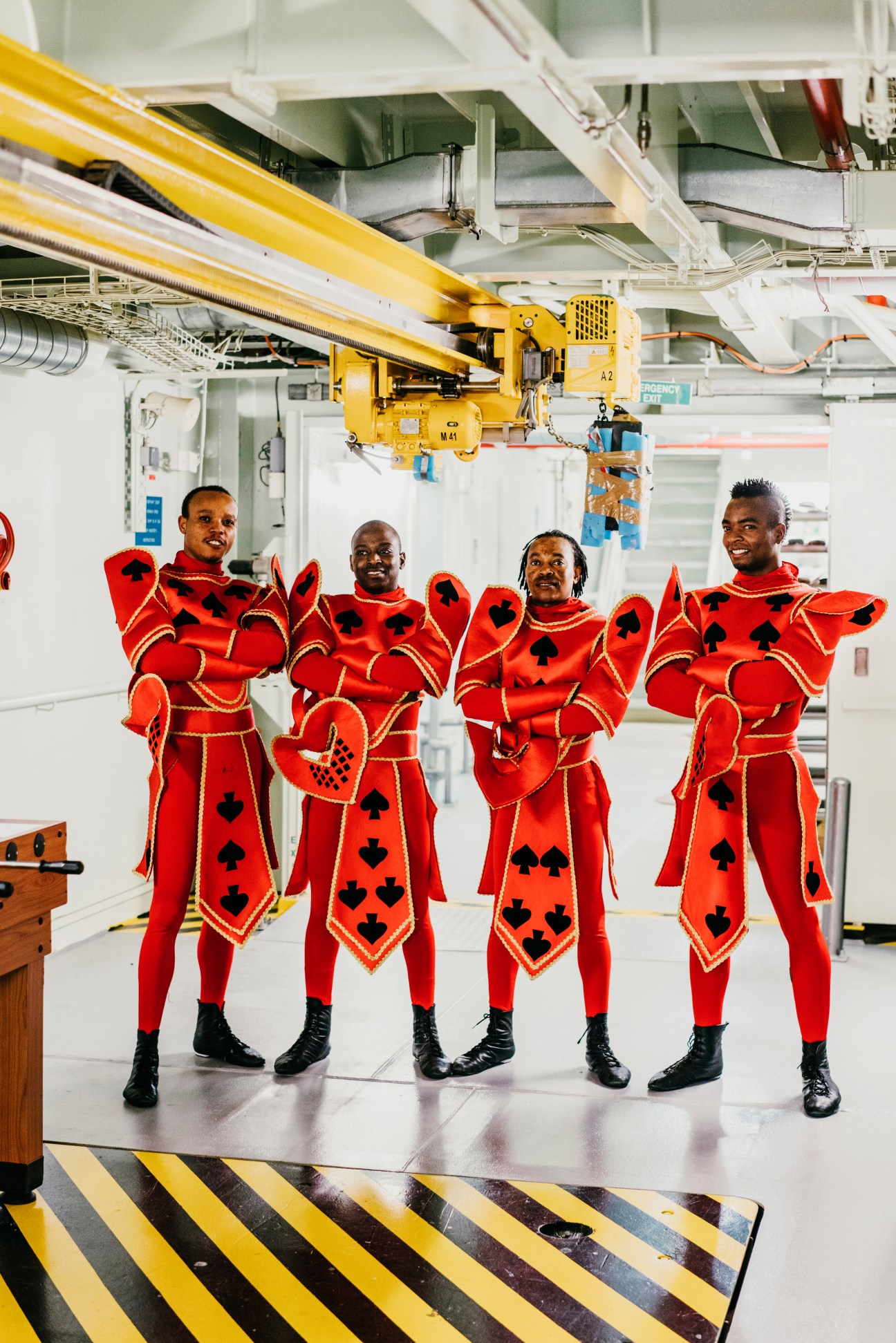 Five performers in red playing card costumes with black suit symbols pose on a ship deck.