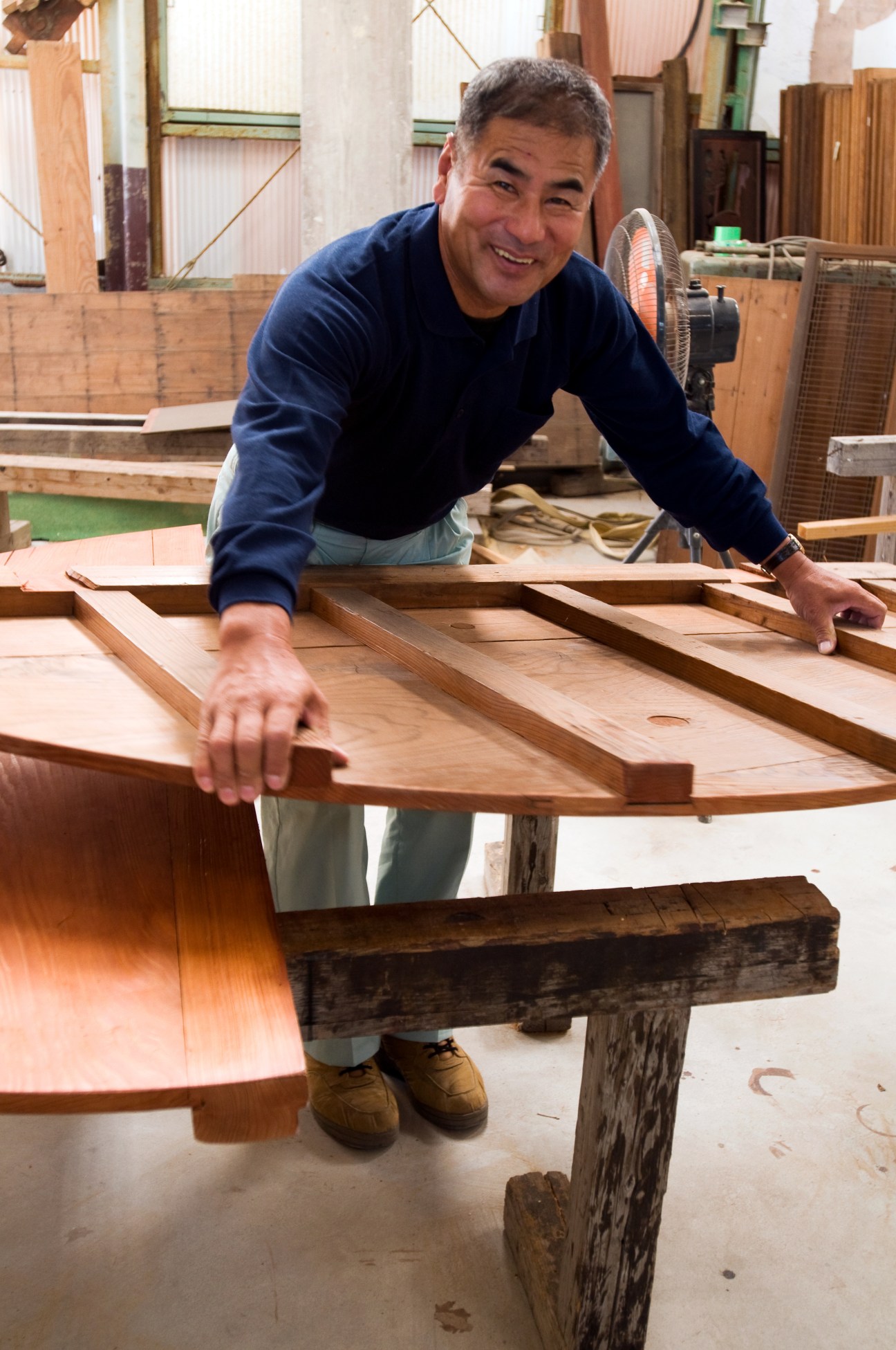 Master carpenter Kazuhiko Fukamura leaning over wooden building components in his workshop.