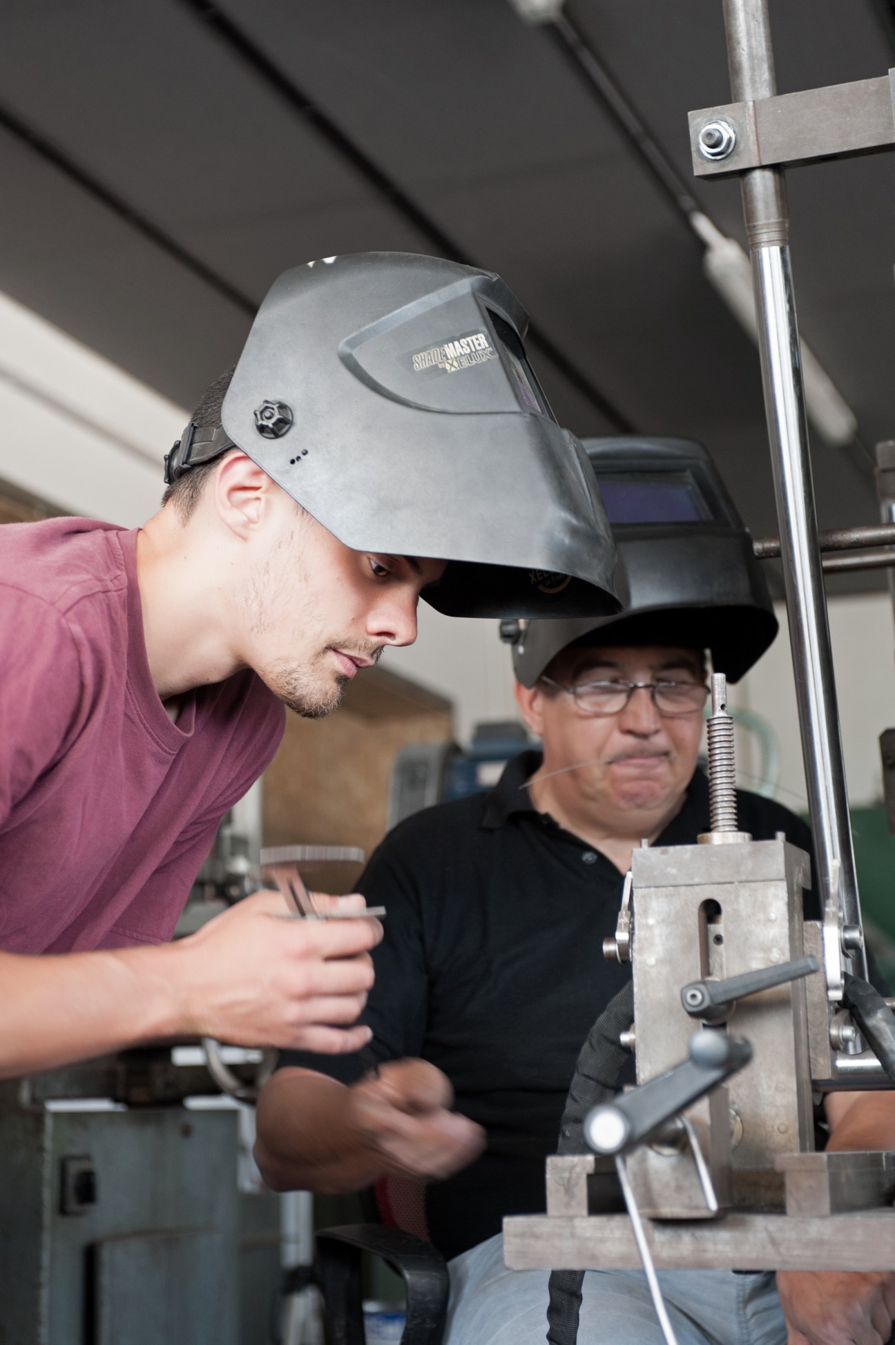 Young apprentice in maroon shirt wearing welding mask observes older craftsman in black shirt preparing stainless-steel tube.