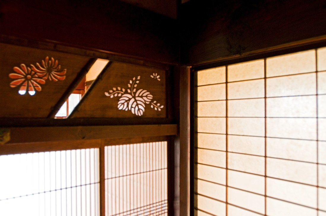 Shoji paper screens with decorative floral cutouts casting shadows inside a traditional Japanese farmhouse interior.