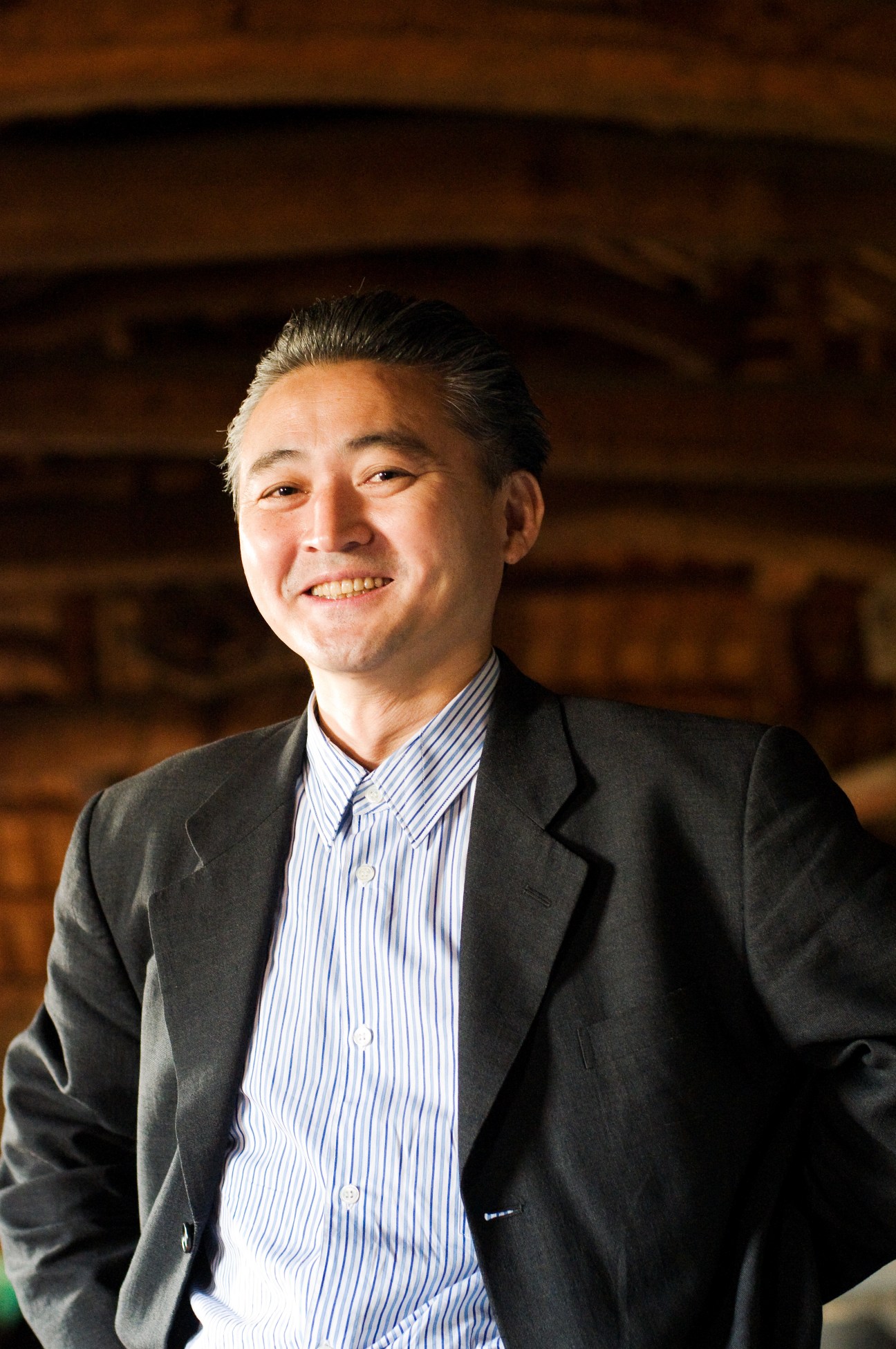 Mikio Shinagawa, a Japanese-American restaurateur, smiles at the camera wearing a dark blazer and blue striped shirt inside a traditional wooden structure.