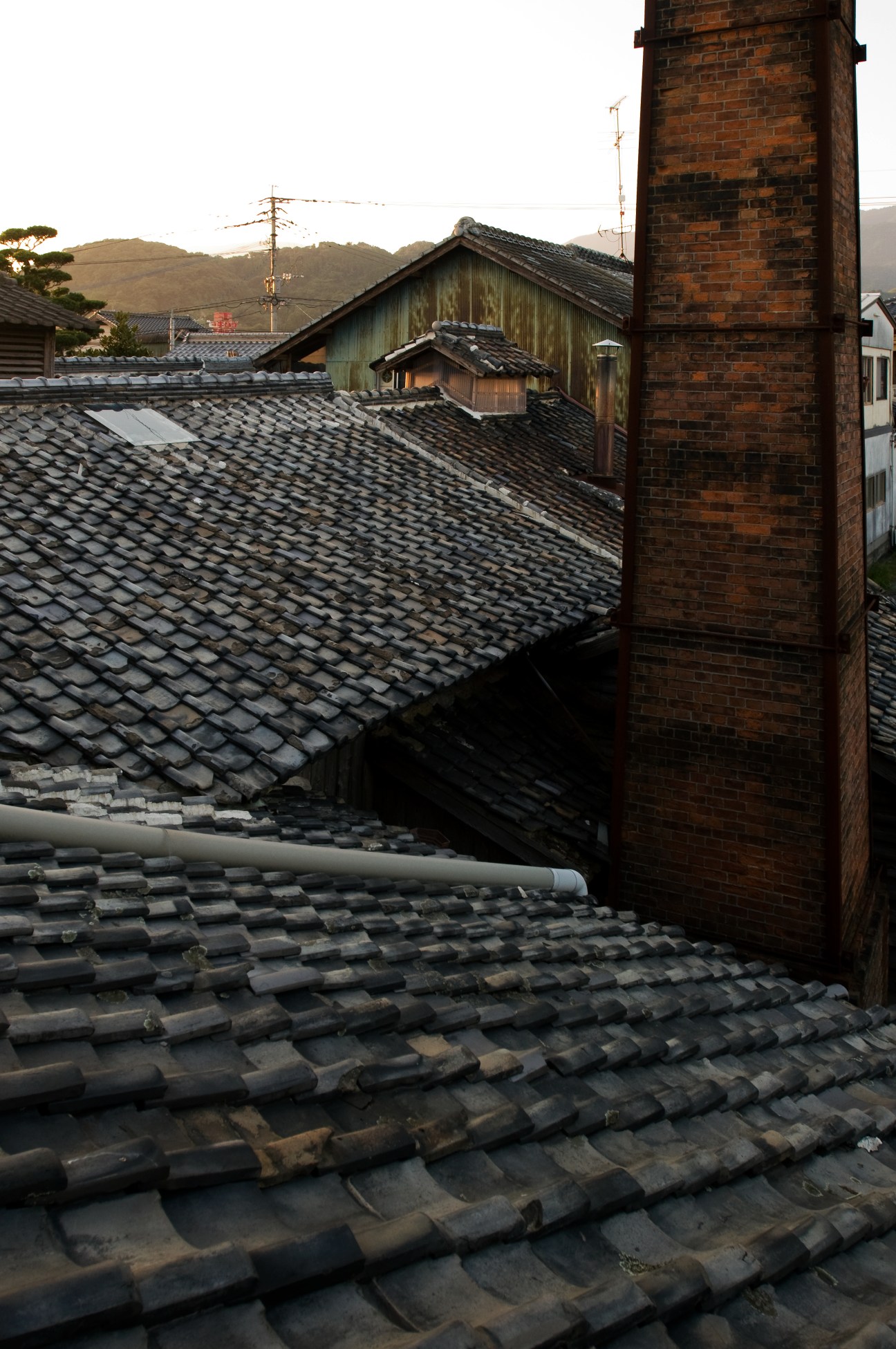 Tiled roof of a traditional Japanese brewery with brick chimney and hillside landscape beyond.