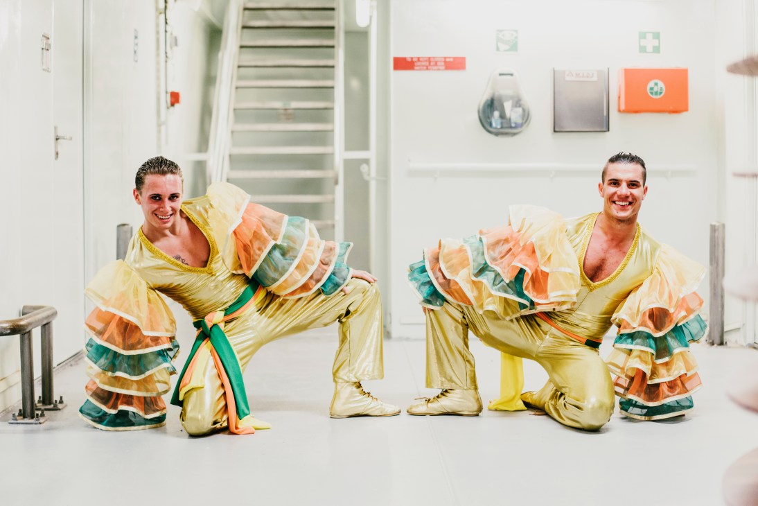 Two male dancers in gold and colorful ruffled costumes pose in a ship corridor.