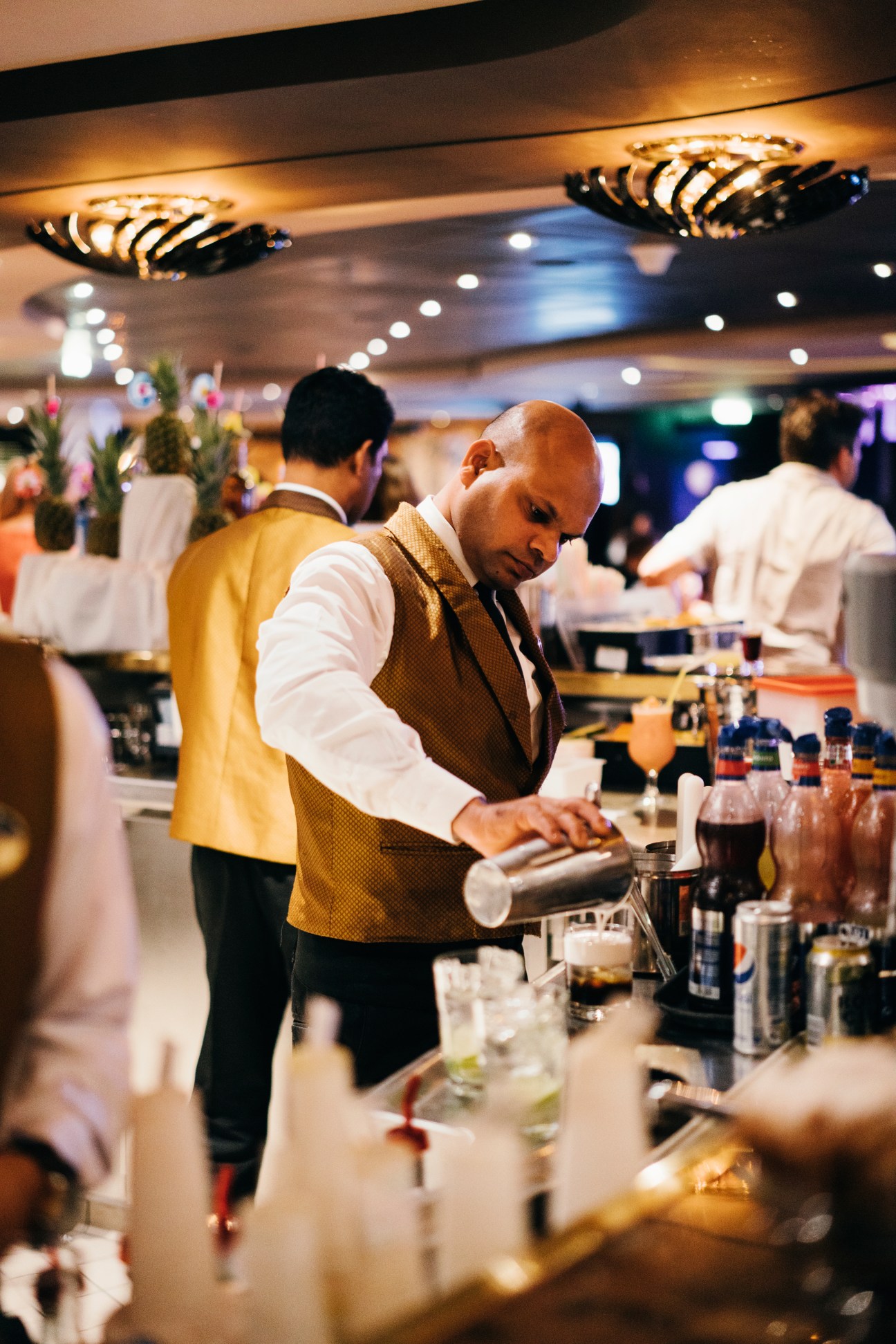 Bartender in gold vest and white shirt pouring a drink at a bar in an elegant lounge with decorative lighting.