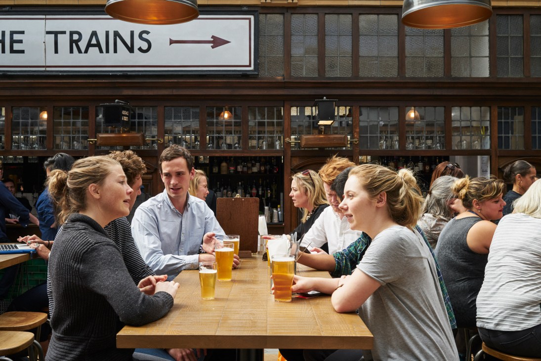 Customers enjoy drinks at a bar counter in a restored railway station with vintage signage reading 'THE TRAINS' overhead.