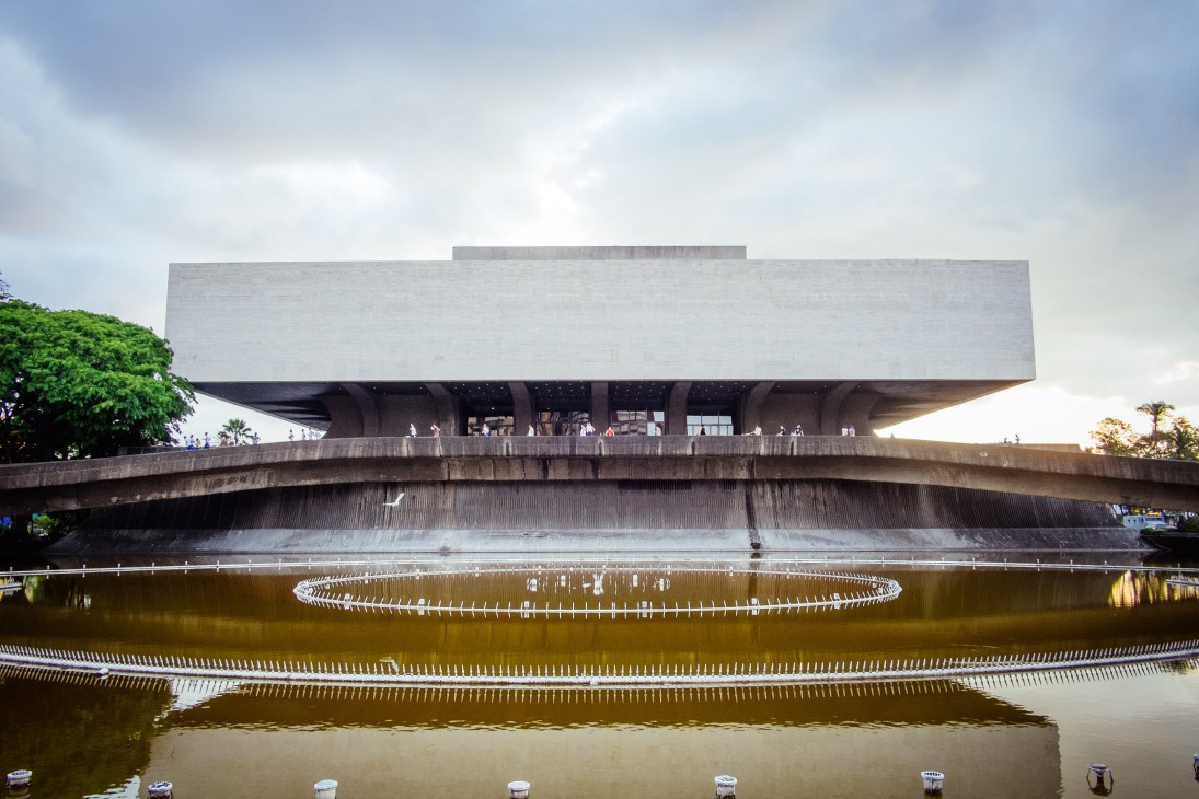 Modernist concrete building with overhanging white upper level reflected in still water below.