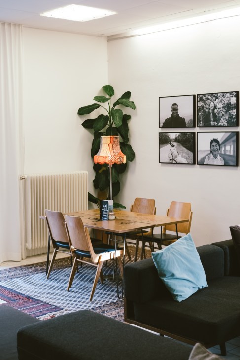 Modern hotel lobby with wooden dining table, mid-century chairs, large potted plant with orange lamp, and black-and-white portrait photographs on white walls.