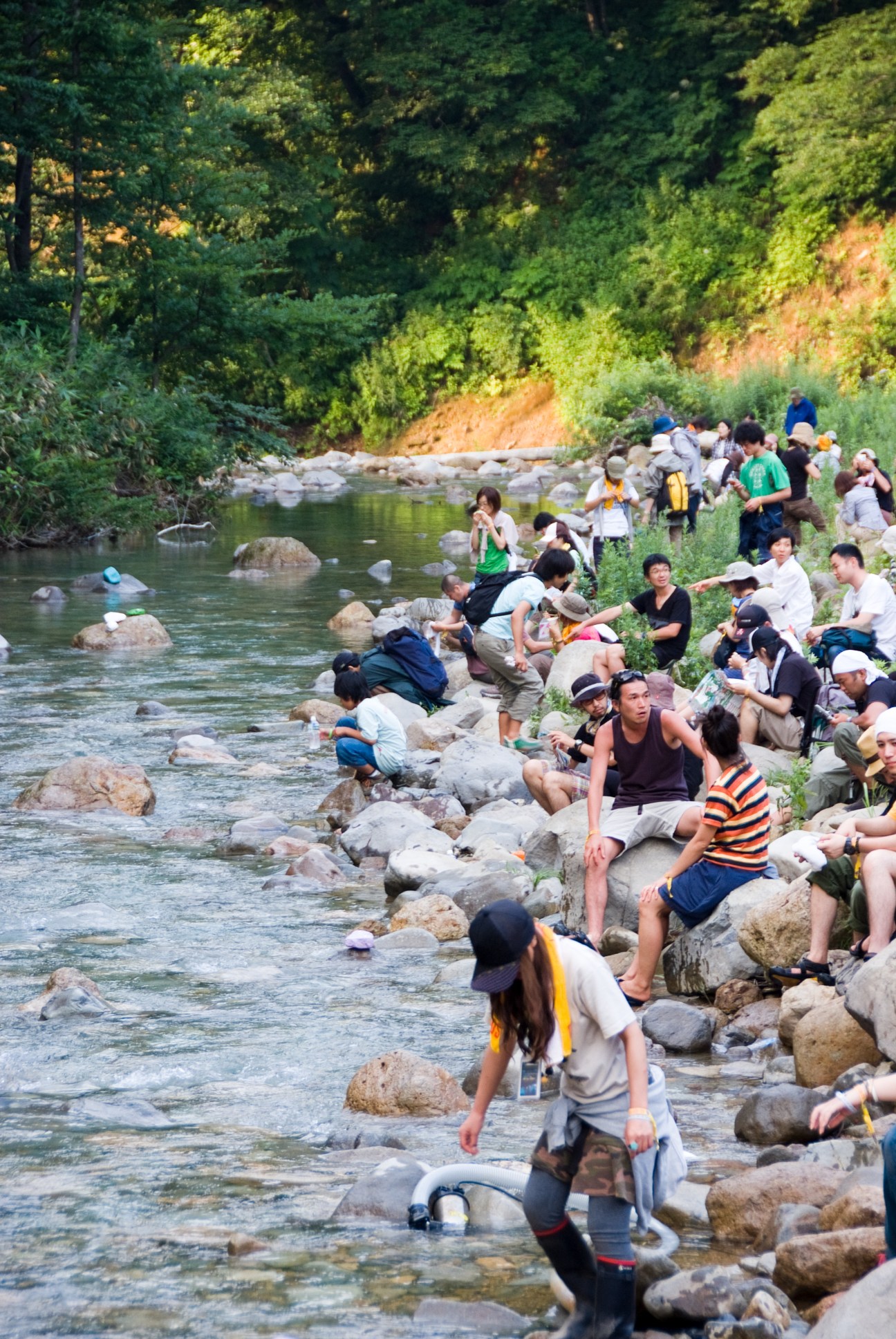Festival attendees relaxing by a river with boulders and forested hillside at Fuji Rock.