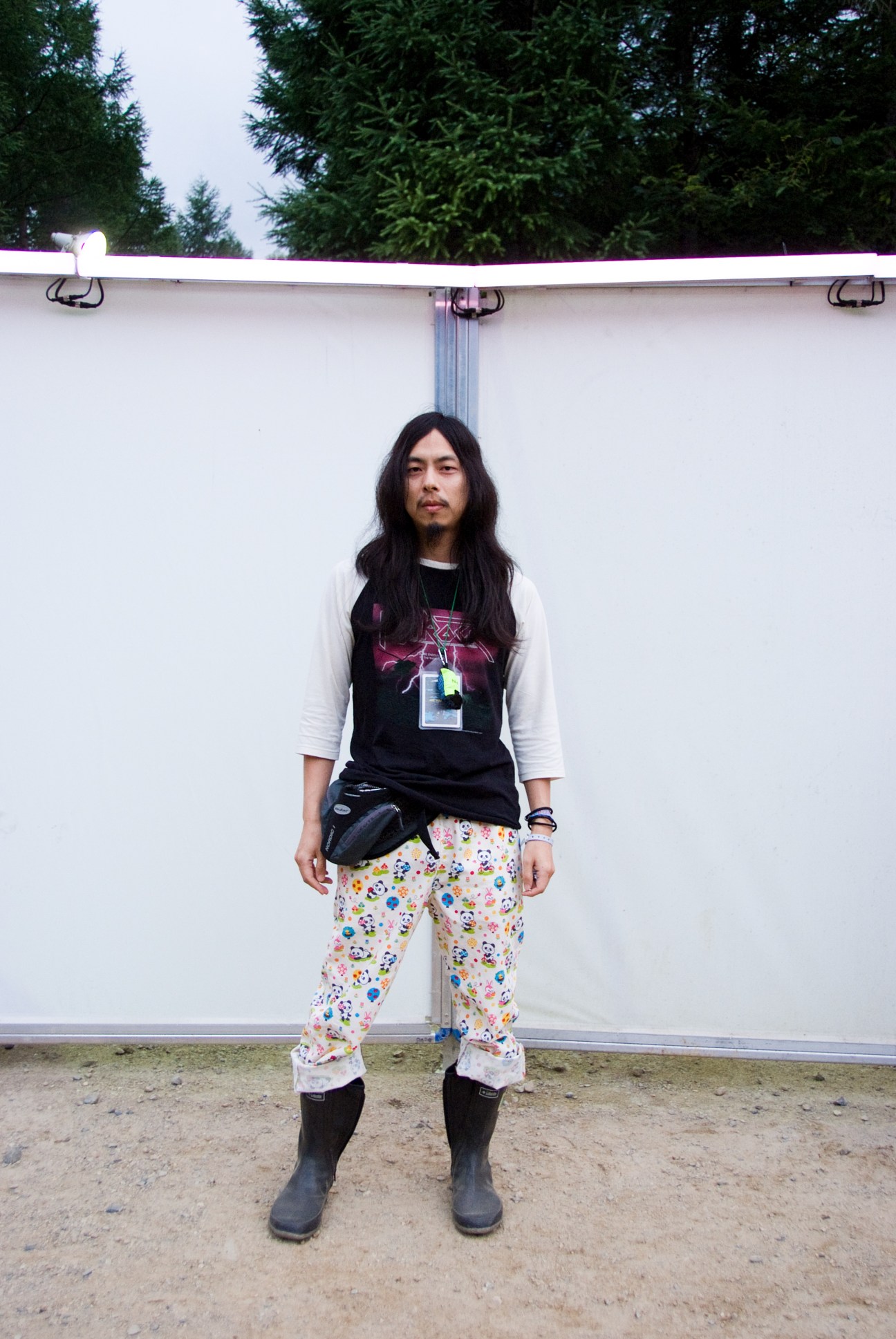 Festival attendee Jun Kumayama wearing graphic band tee, patterned pants, and black boots against white backdrop.