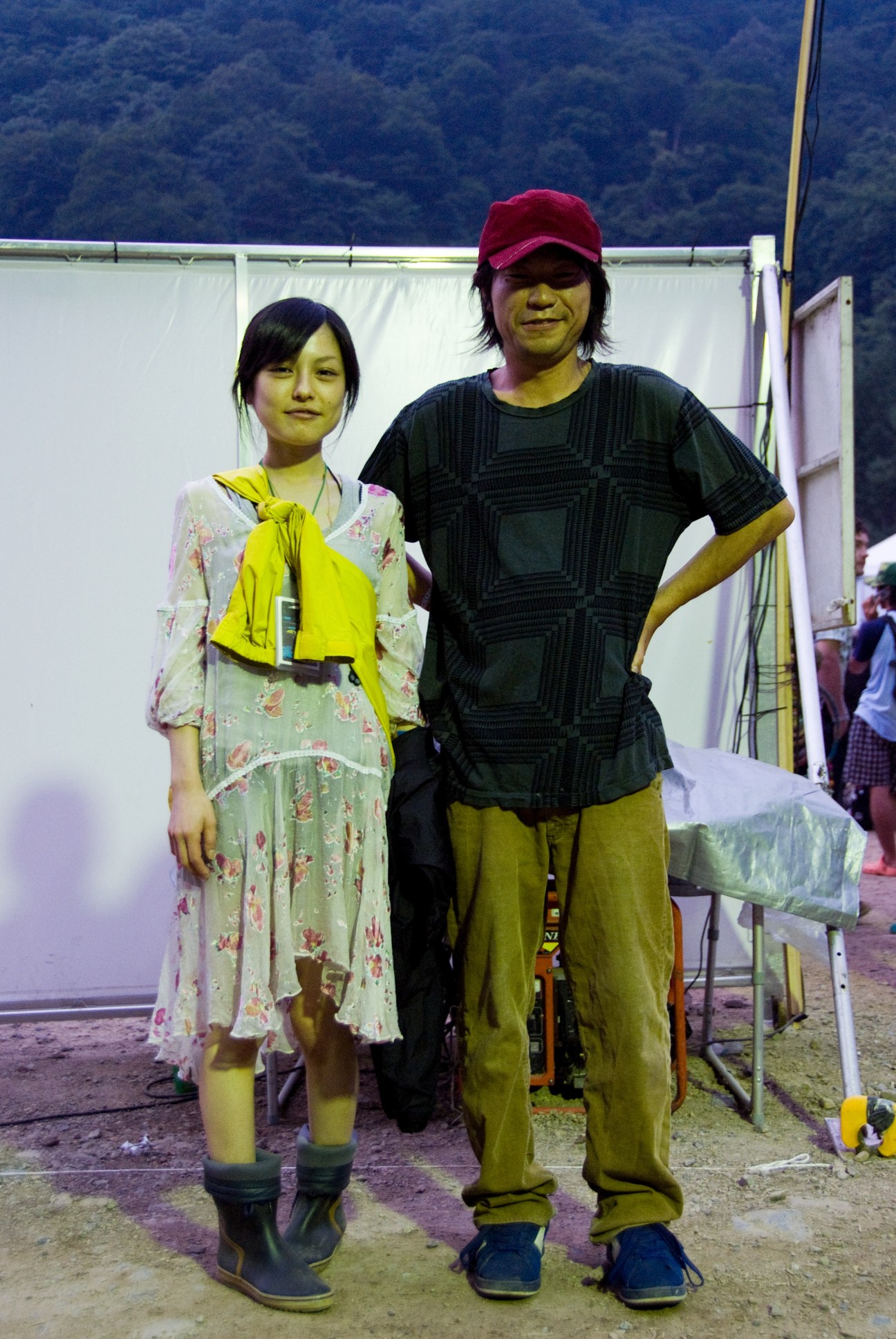 Two festival-goers pose together at Fuji Rock, wearing colorful casual clothing and boots against a backdrop of white tent panels and forested hills.