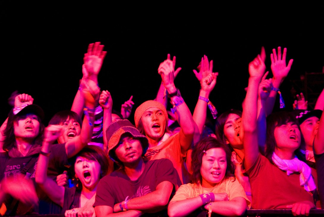 Enthusiastic crowd of Japanese festival-goers with raised arms and excited expressions bathed in pink stage lighting.