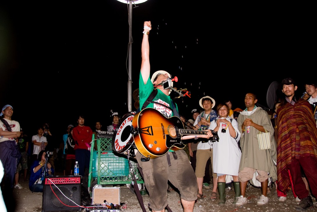 A one-man band performer wearing sunglasses and green shirt plays guitar to a crowd standing in a queue at night.
