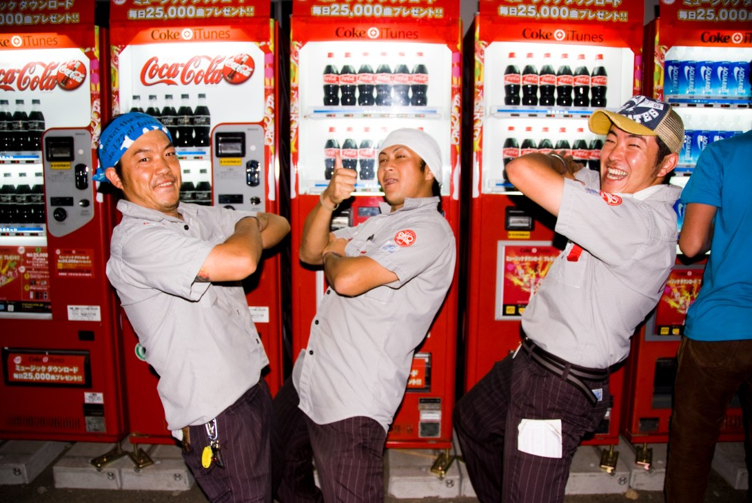 Three uniformed Coca-Cola vending machine service staff pose cheerfully in front of red Coke vending machines at Fuji Rock festival.