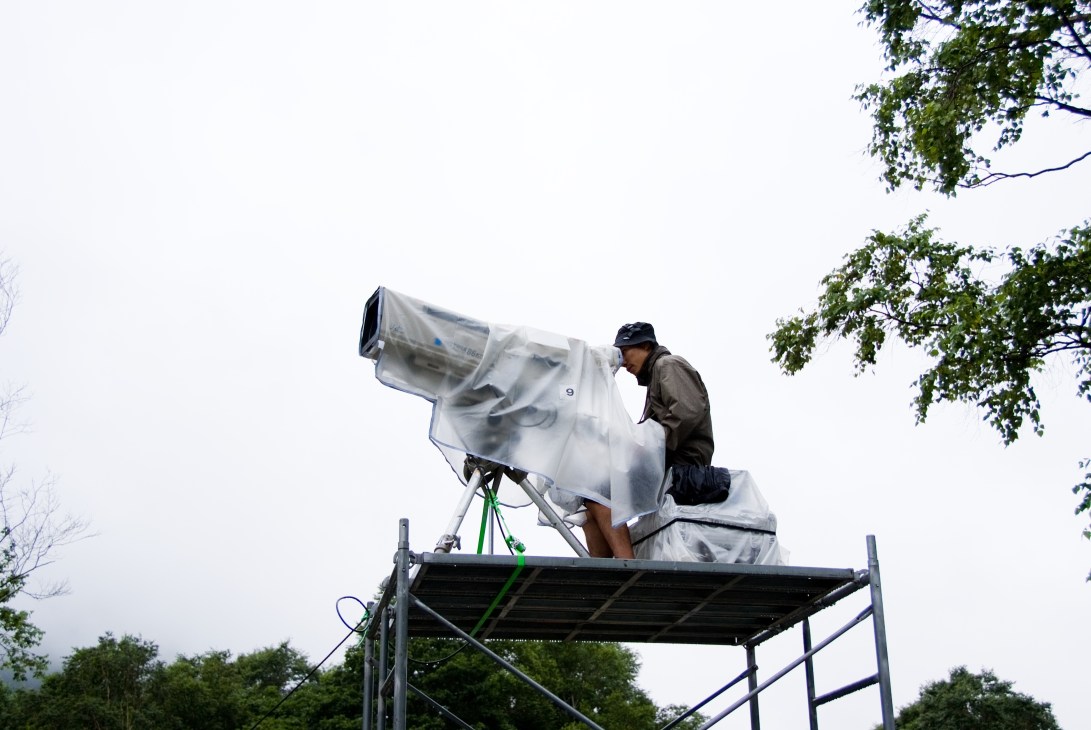 A cameraman on an elevated platform covers the festival with a large television camera wrapped in protective plastic.