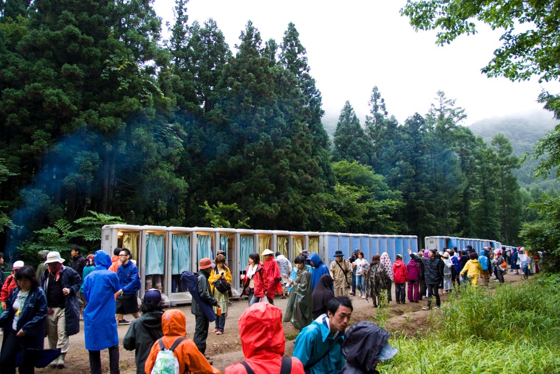 Festival-goers queue at portable toilets surrounded by forested mountains at Fuji Rock.