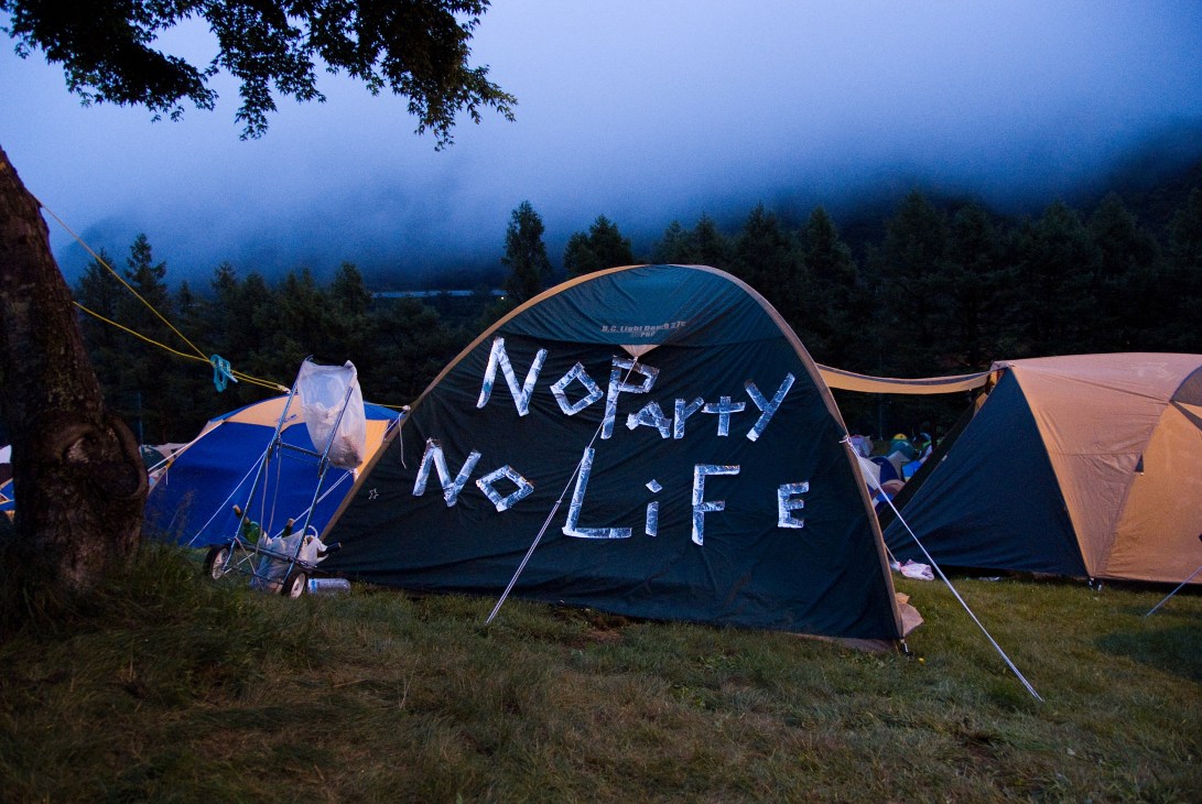 Festival campers' tents at dusk with 'No Party No Life' written on dark tent among colorful ones in misty mountain landscape.