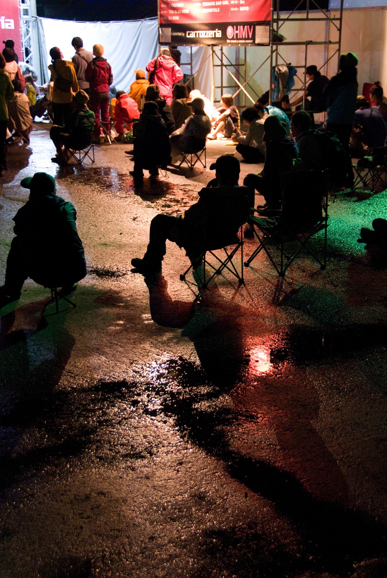 Festival-goers sitting in camping chairs wait in a wet outdoor queue at night, with colorful tent structures and stage lighting visible in the background.