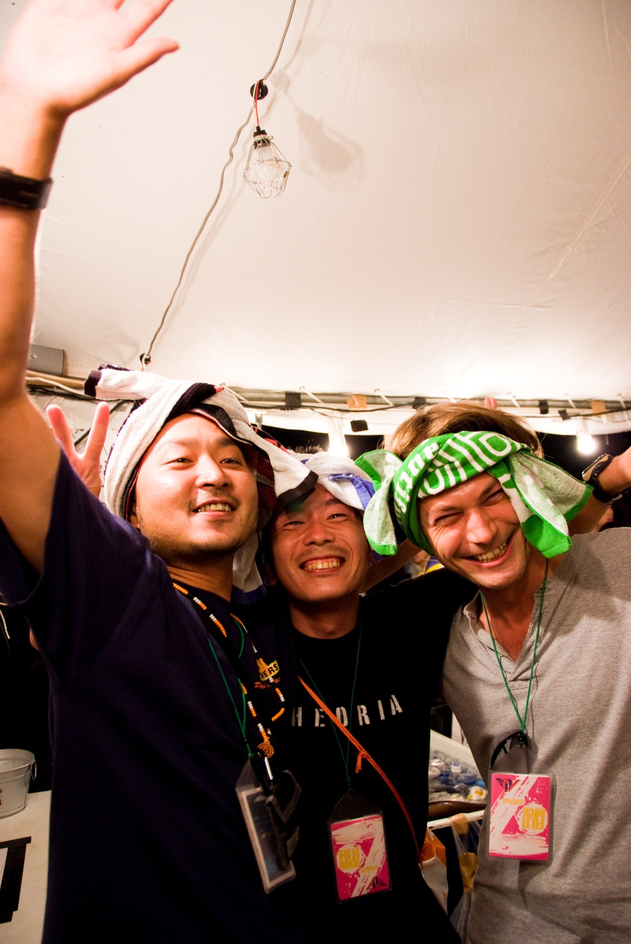 Three smiling festival-goers wearing towels tied as headbands and scarves inside a tent at Fuji Rock Festival.