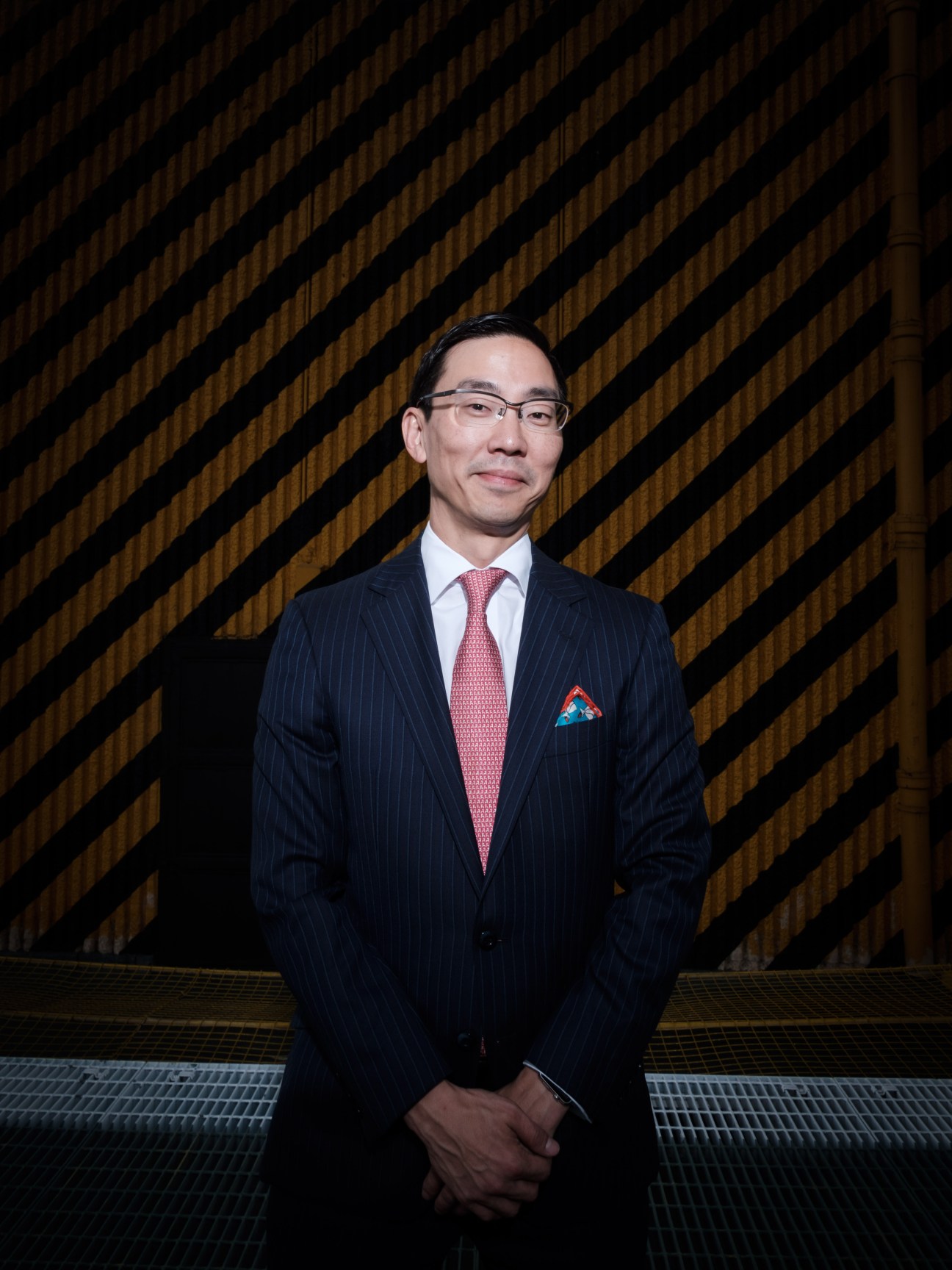 Man in navy pinstriped suit with glasses and red tie standing against diagonal wooden slat background.