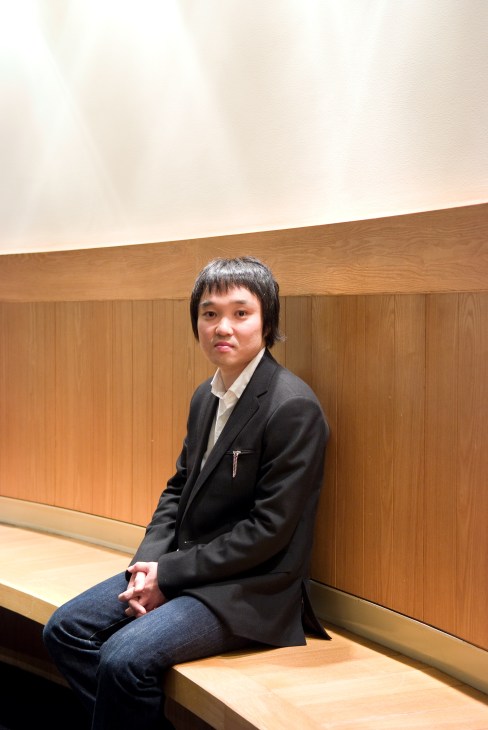 Hiroaki Narita, founder of Connect furniture auction house, sitting on a wooden bench against a wood-paneled wall.